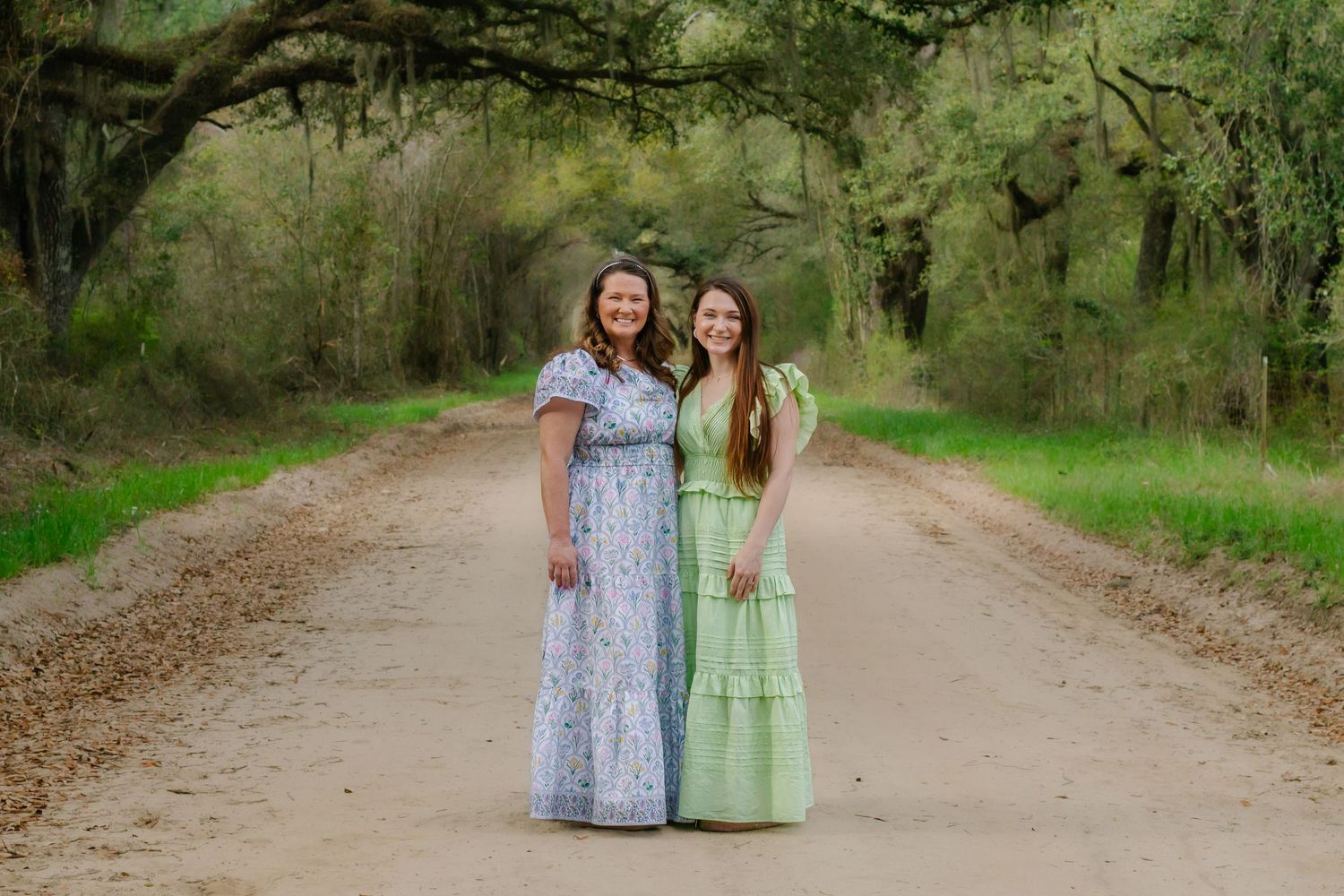 Two women in maxi dresses smile together on a tree-lined dirt road.