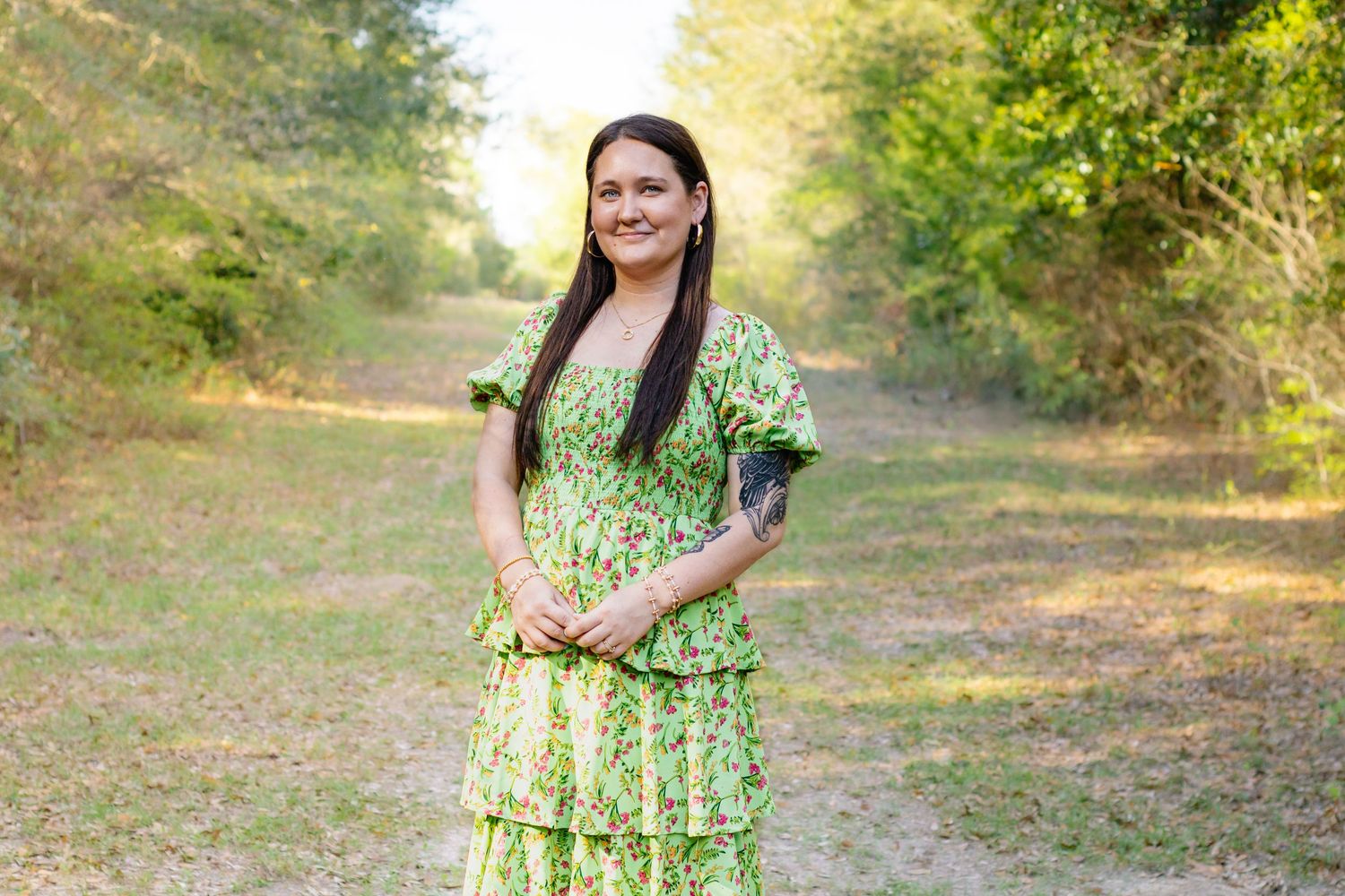 Smiling woman in green floral dress standing on a wooded dirt path in natural sunlight.