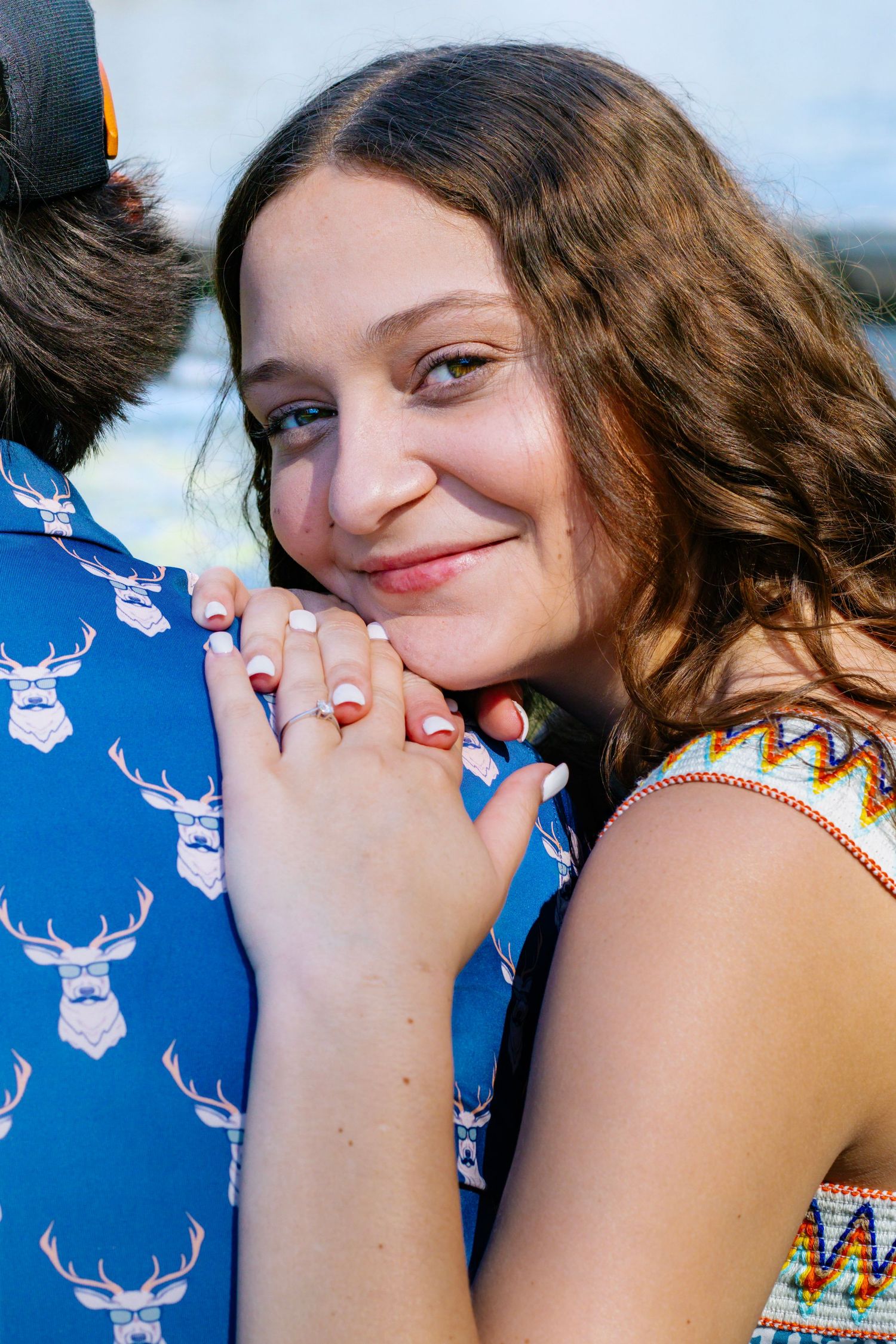 Young woman with curly brown hair smiles while embracing man in blue deer print shirt, showing engagement ring.