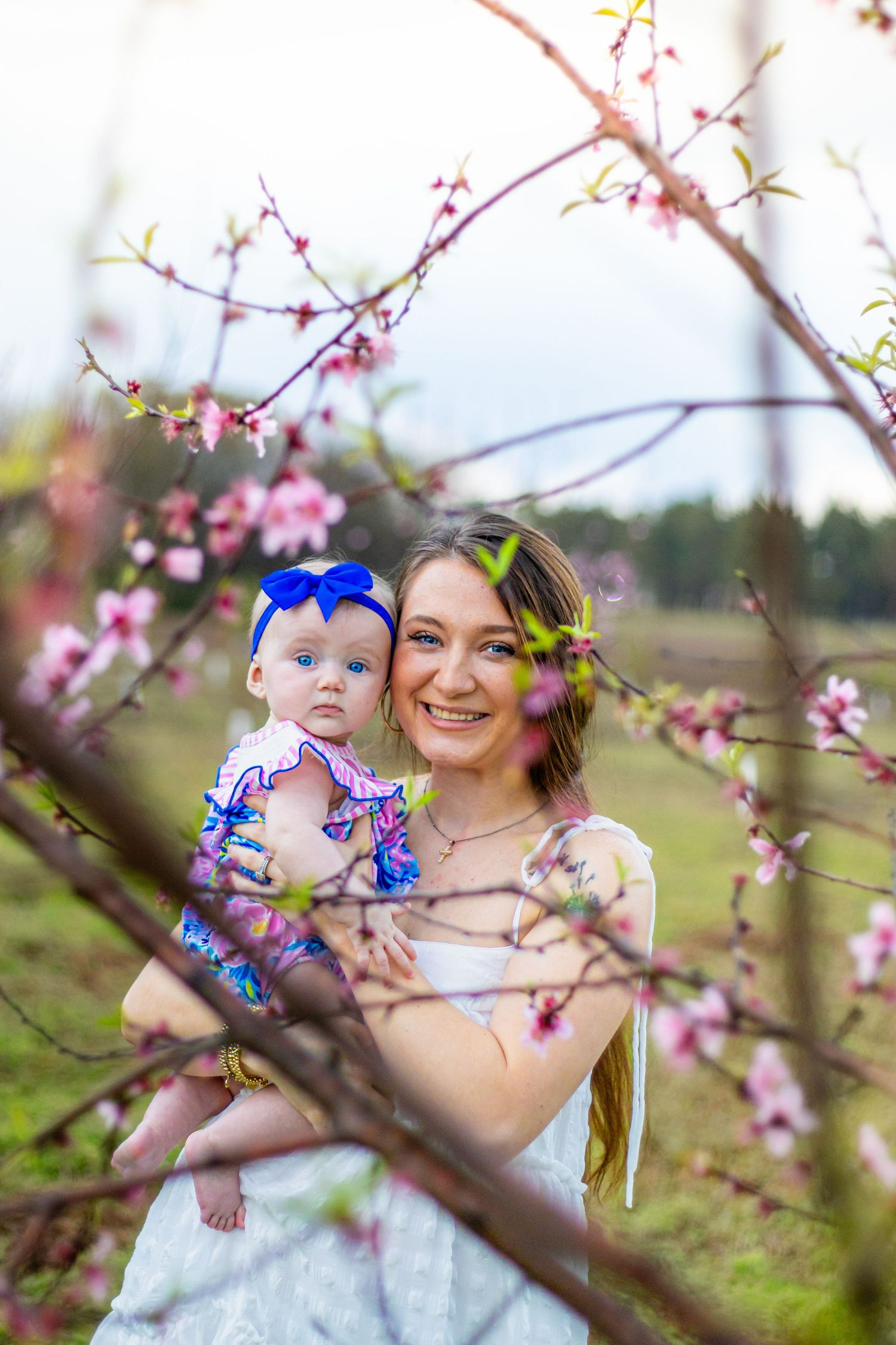 Smiling young mother in white dress holds baby girl in floral outfit among pink blossoming tree branches outdoors.
