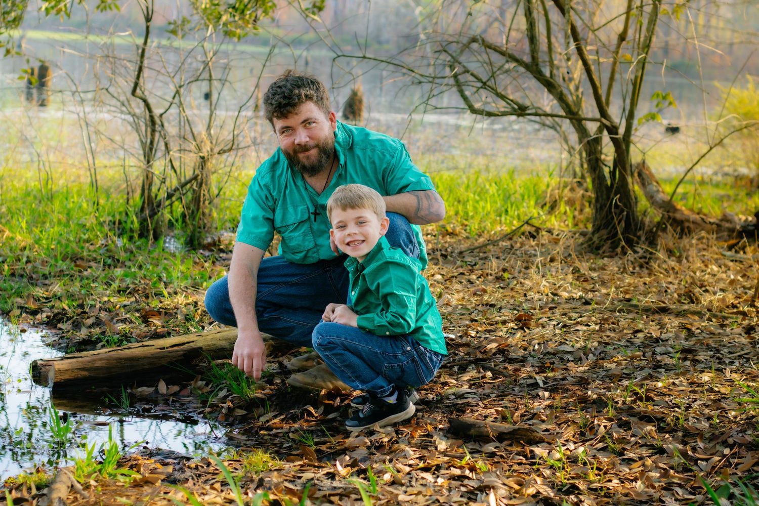 Father and young son in green shirts crouch together near a small stream in autumn woods.