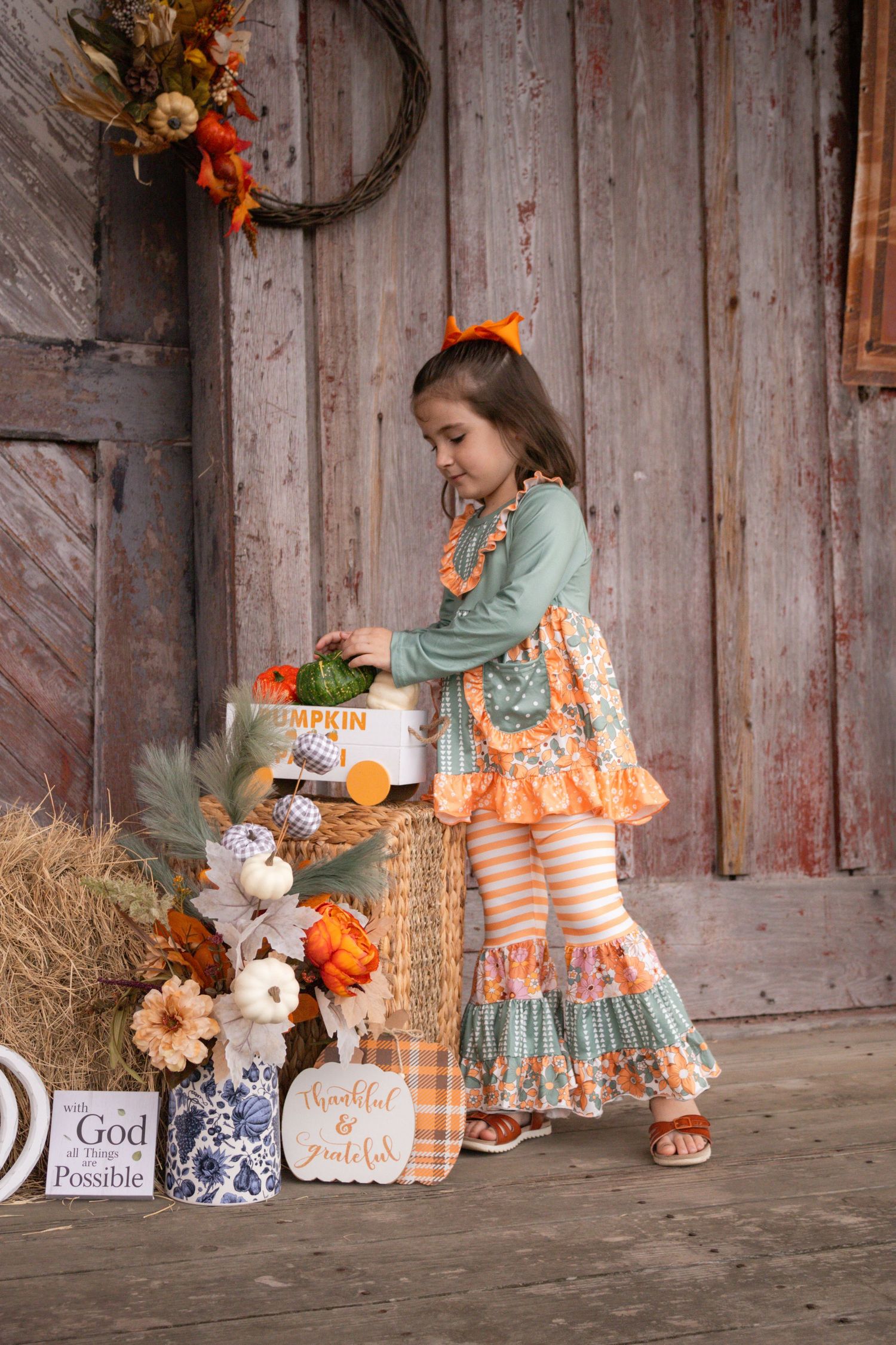 Young girl in festive fall outfit reaches into pumpkin basket beside hay bales and autumn decorations.
