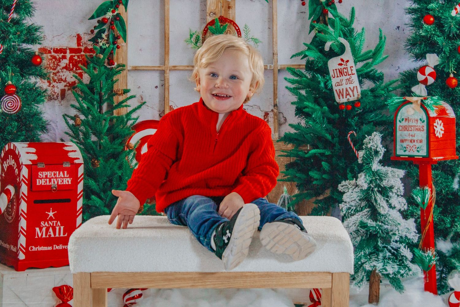 A cheerful blond toddler in a red zip-up sweater sits on a white bench in a Christmas-themed photo studio.