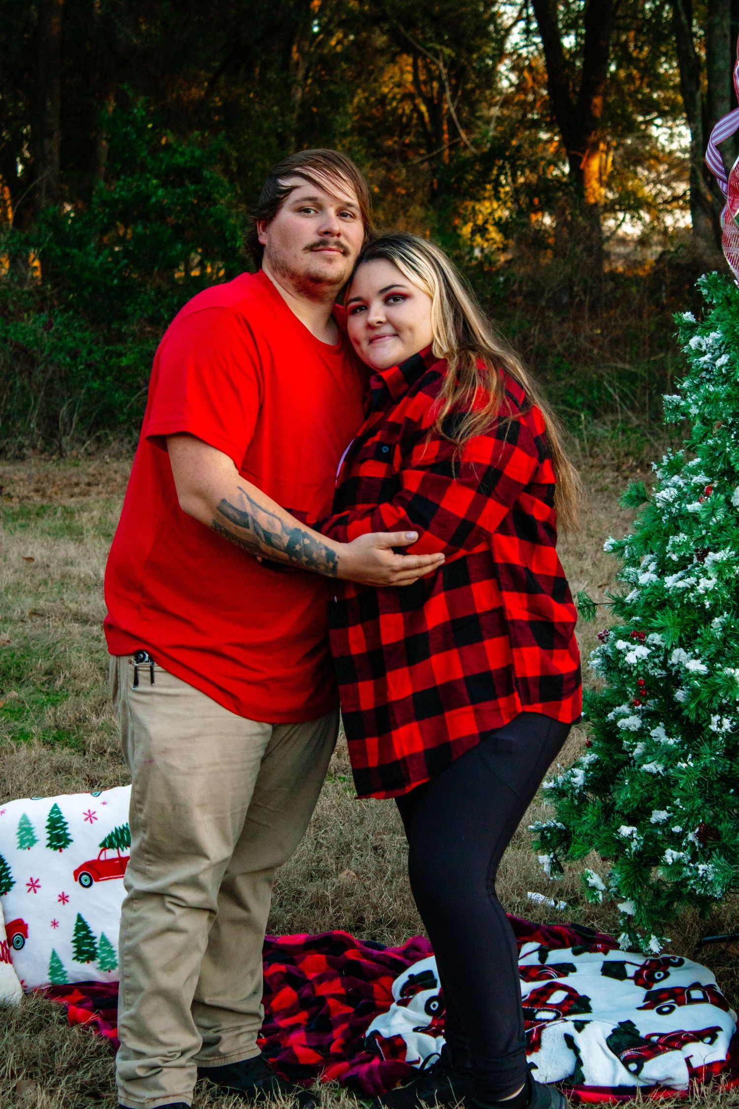 Couple in red outfits embracing near a snowy Christmas tree outdoors, with the woman cradling her baby bump.