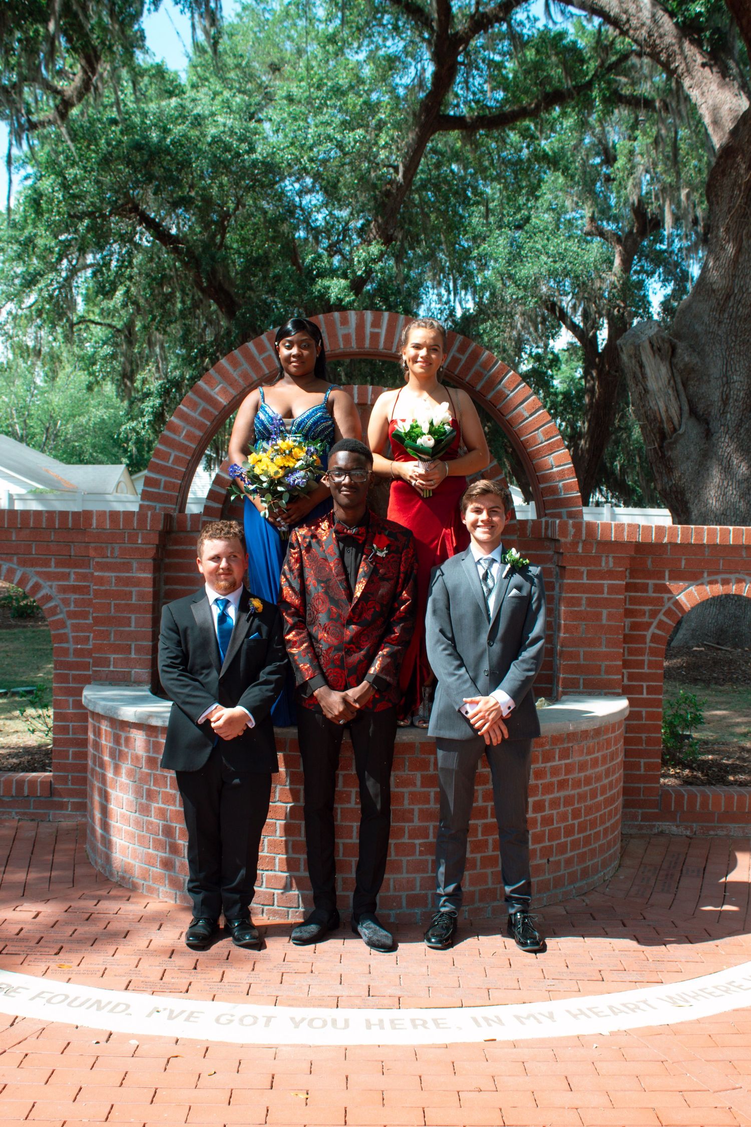Five prom attendees pose at a brick arch monument, girls in formal gowns holding bouquets, guys in suits.