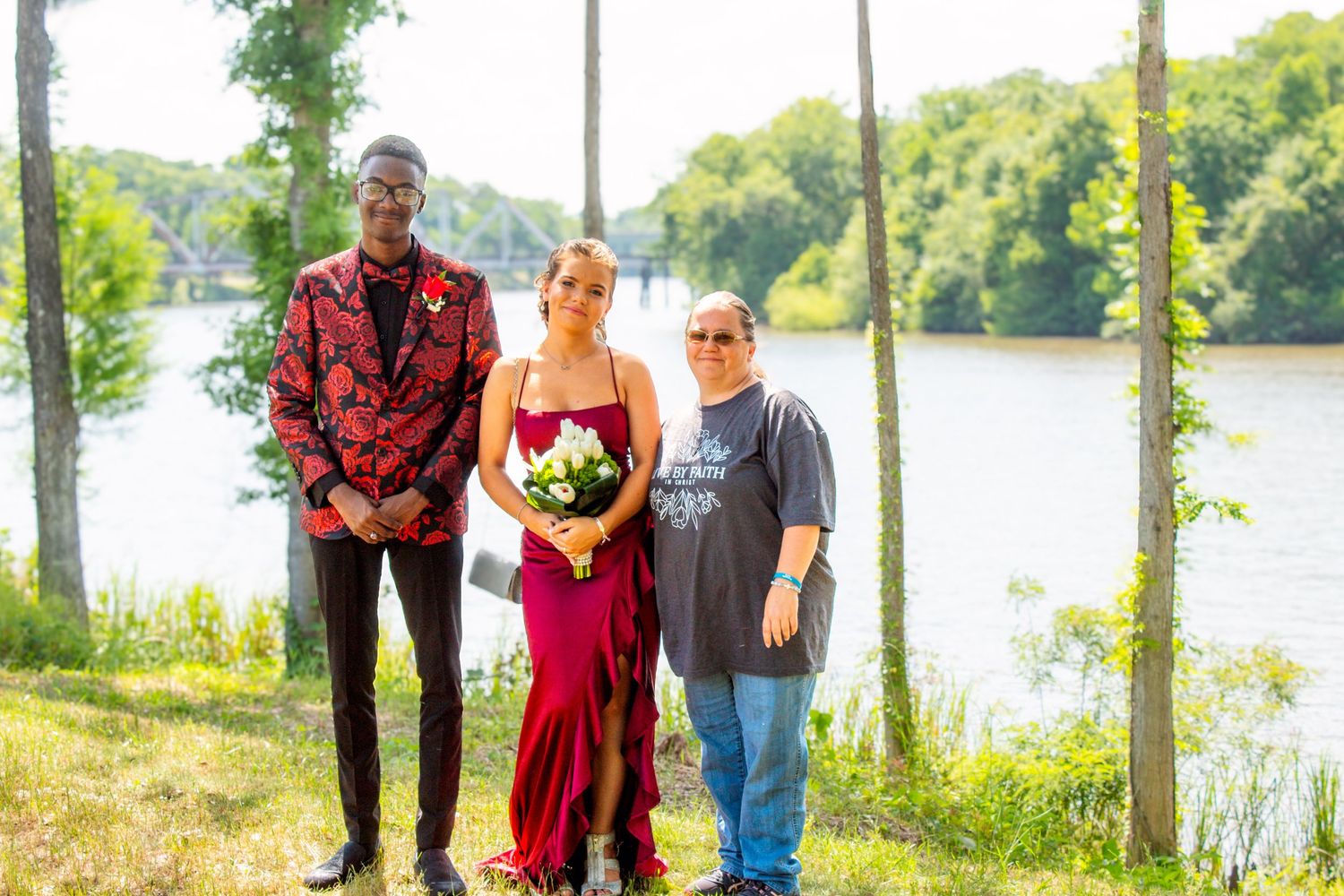 A tall young man in red floral jacket stands with a woman in red gown and older adult outdoors.