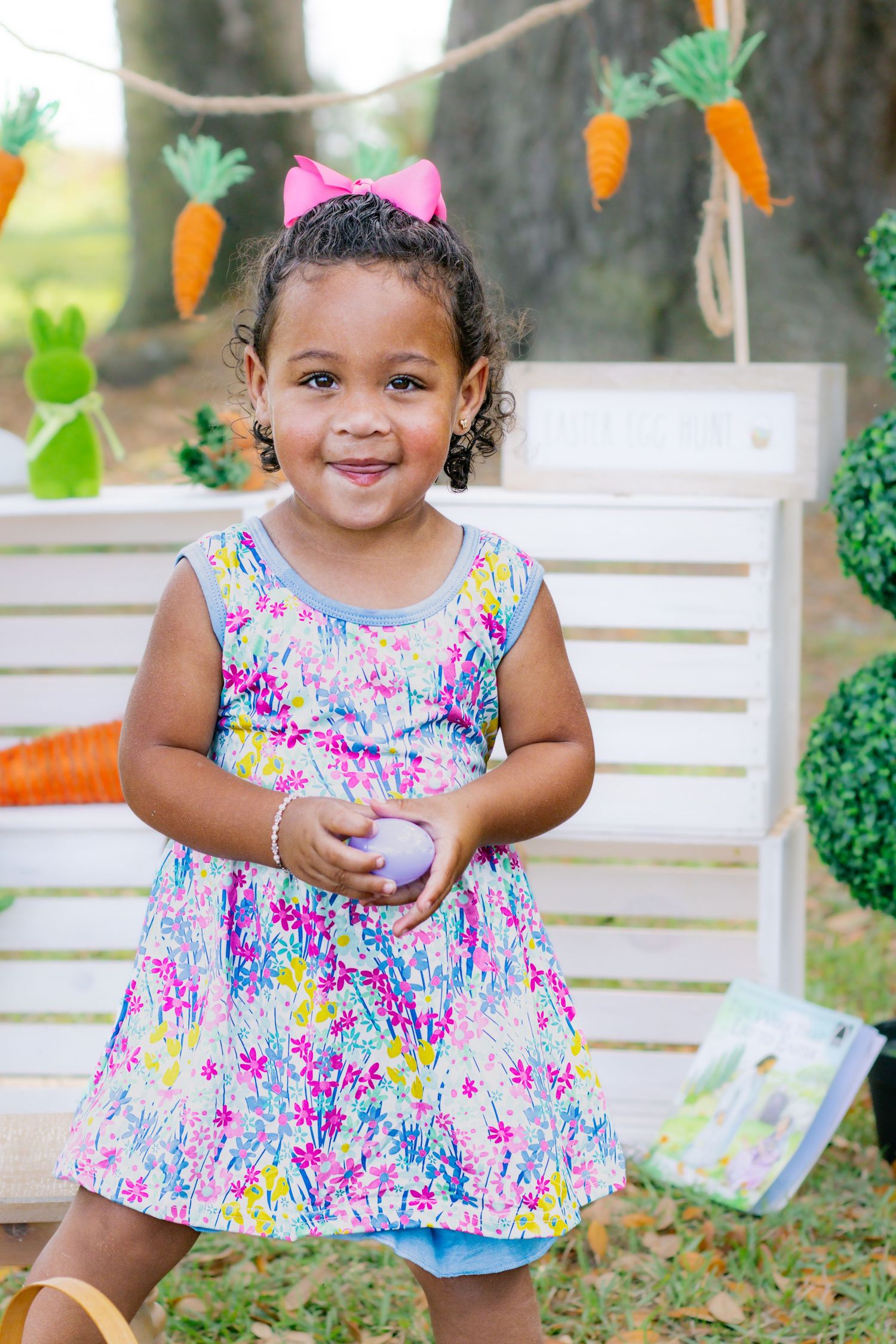 Smiling toddler girl in floral dress holds Easter egg at outdoor spring photo session.