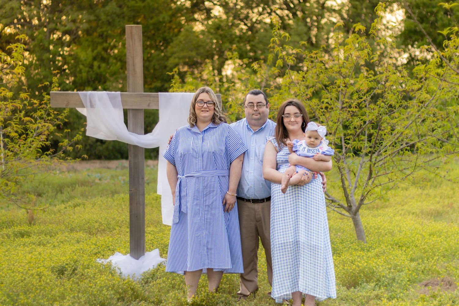 Family of four posing beside a wooden cross with white cloth draped over it in a sunny meadow.