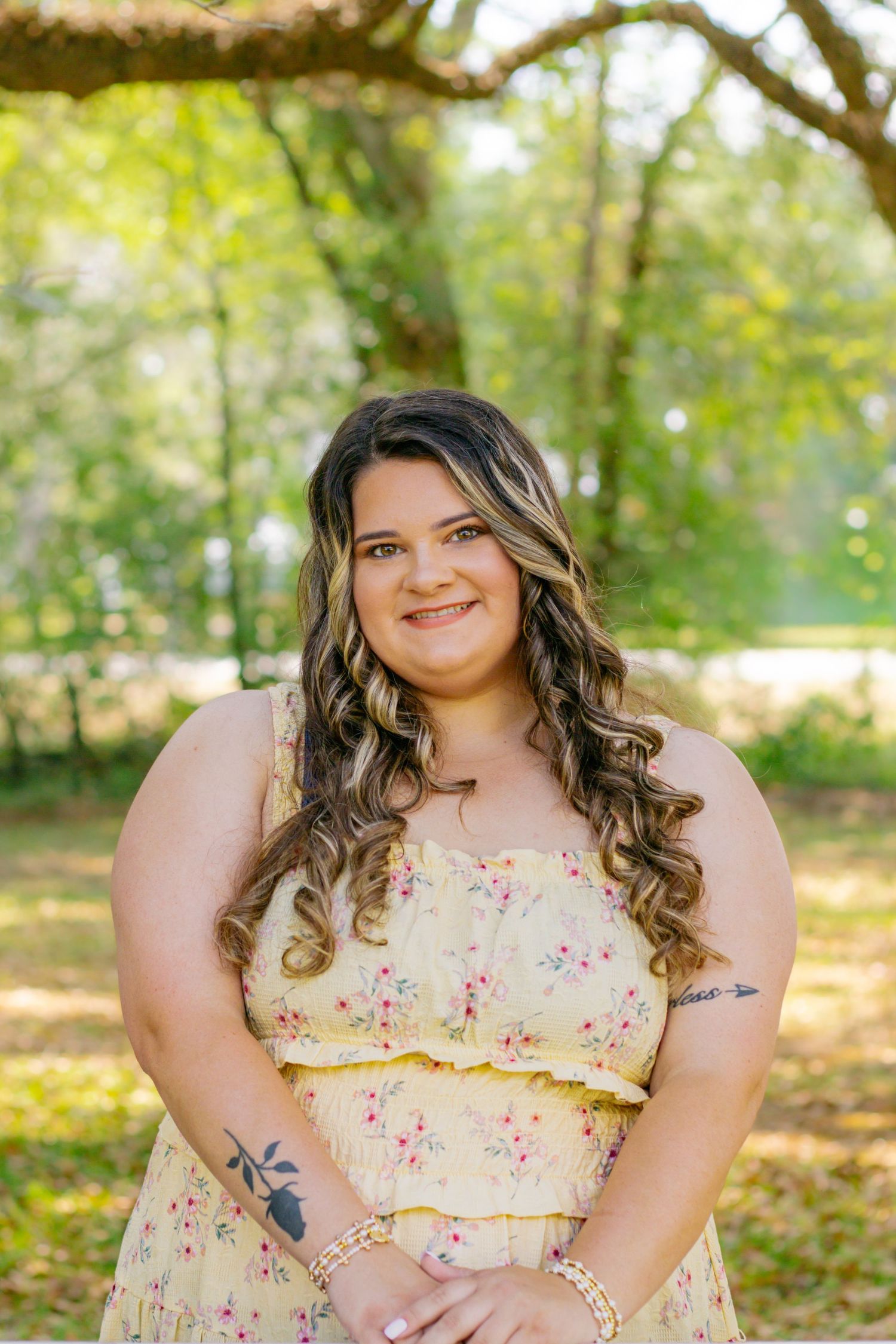 Young woman with curly hair smiles in yellow floral sundress with tattoos in sunny park setting.