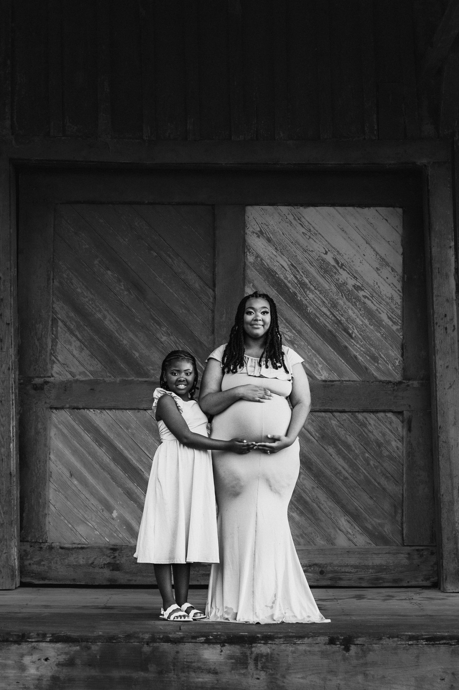 Black and white photo of pregnant woman in white gown with young girl hugging her baby bump.