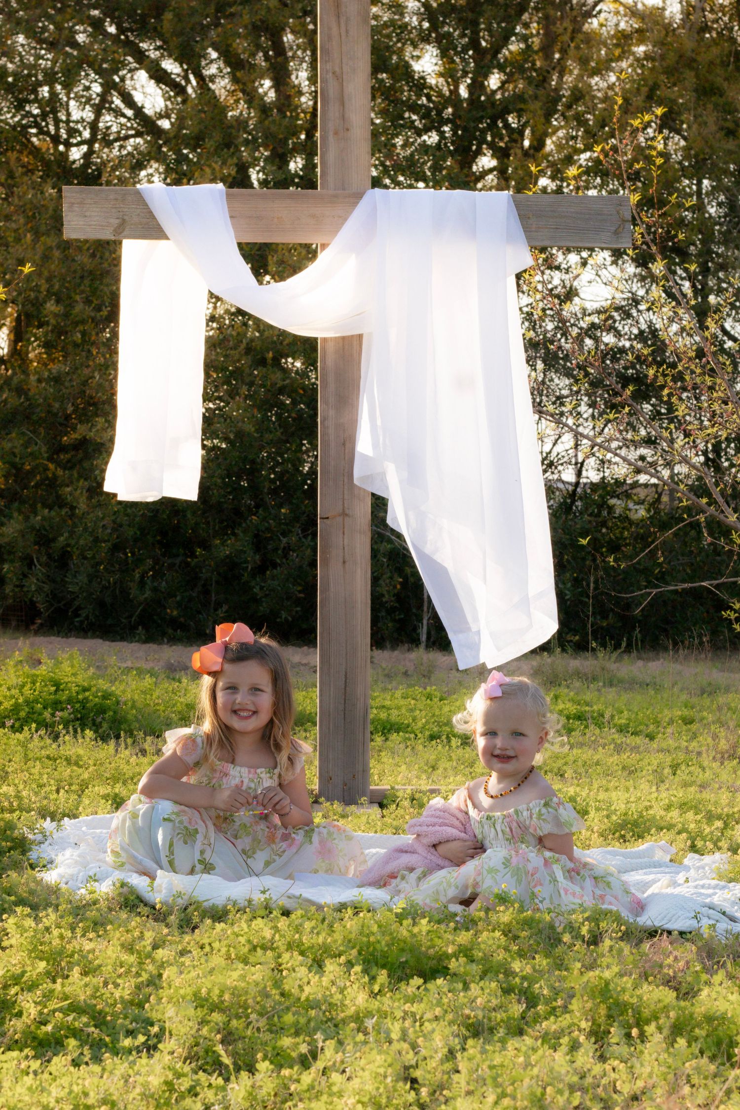 Two young girls in floral dresses smiling beside a wooden cross with white drape in a field.