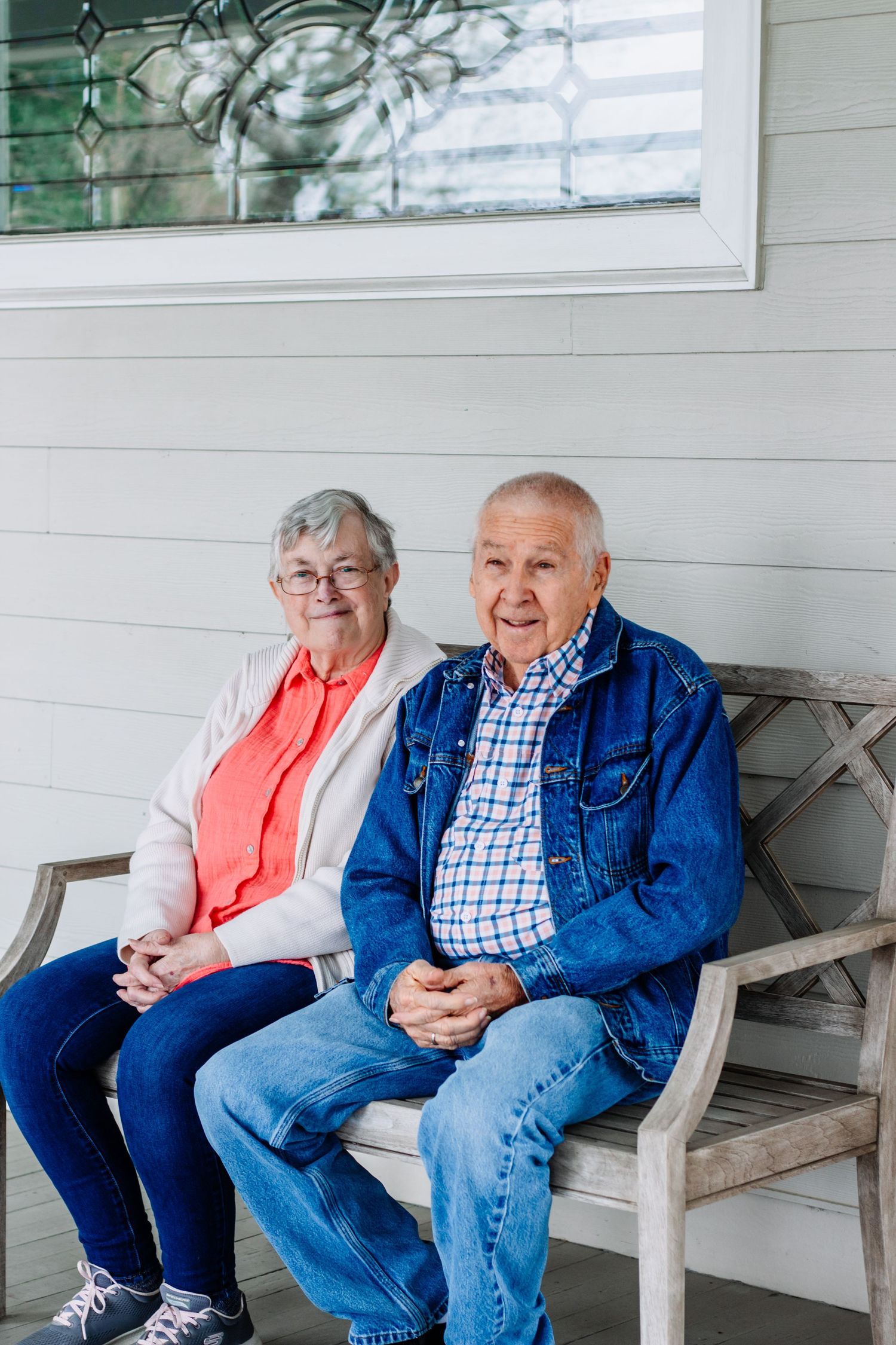 Elderly couple seated on a wooden bench on a porch, smiling at camera.