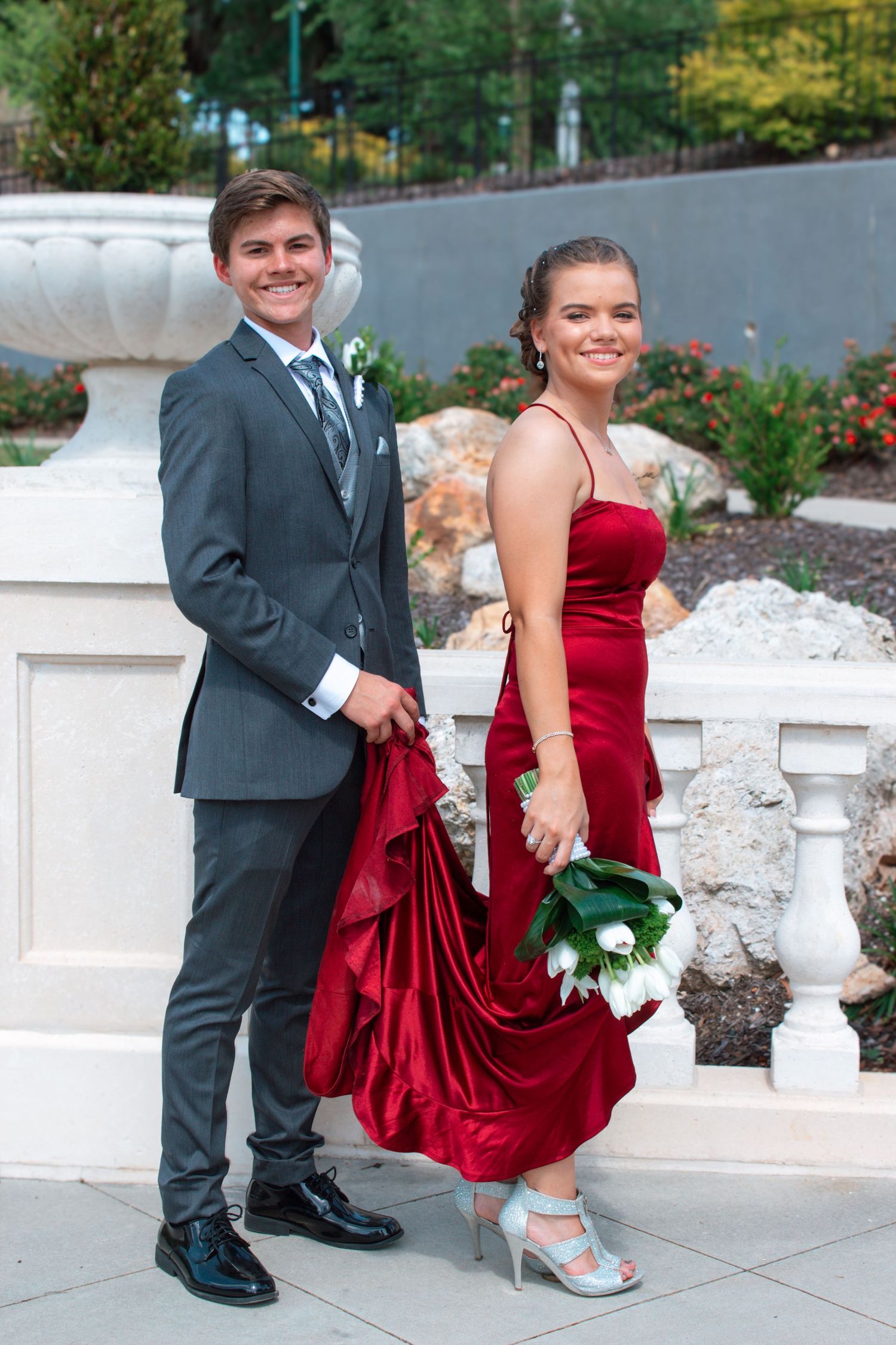 A smiling teenage couple in formal prom attire, boy in gray suit and girl in red gown holding bouquet outdoors.