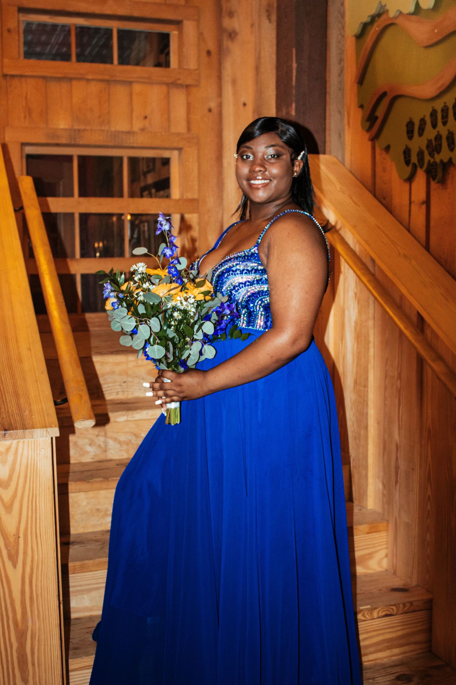 Young woman in a royal blue formal gown holding a yellow and blue floral bouquet on a staircase.