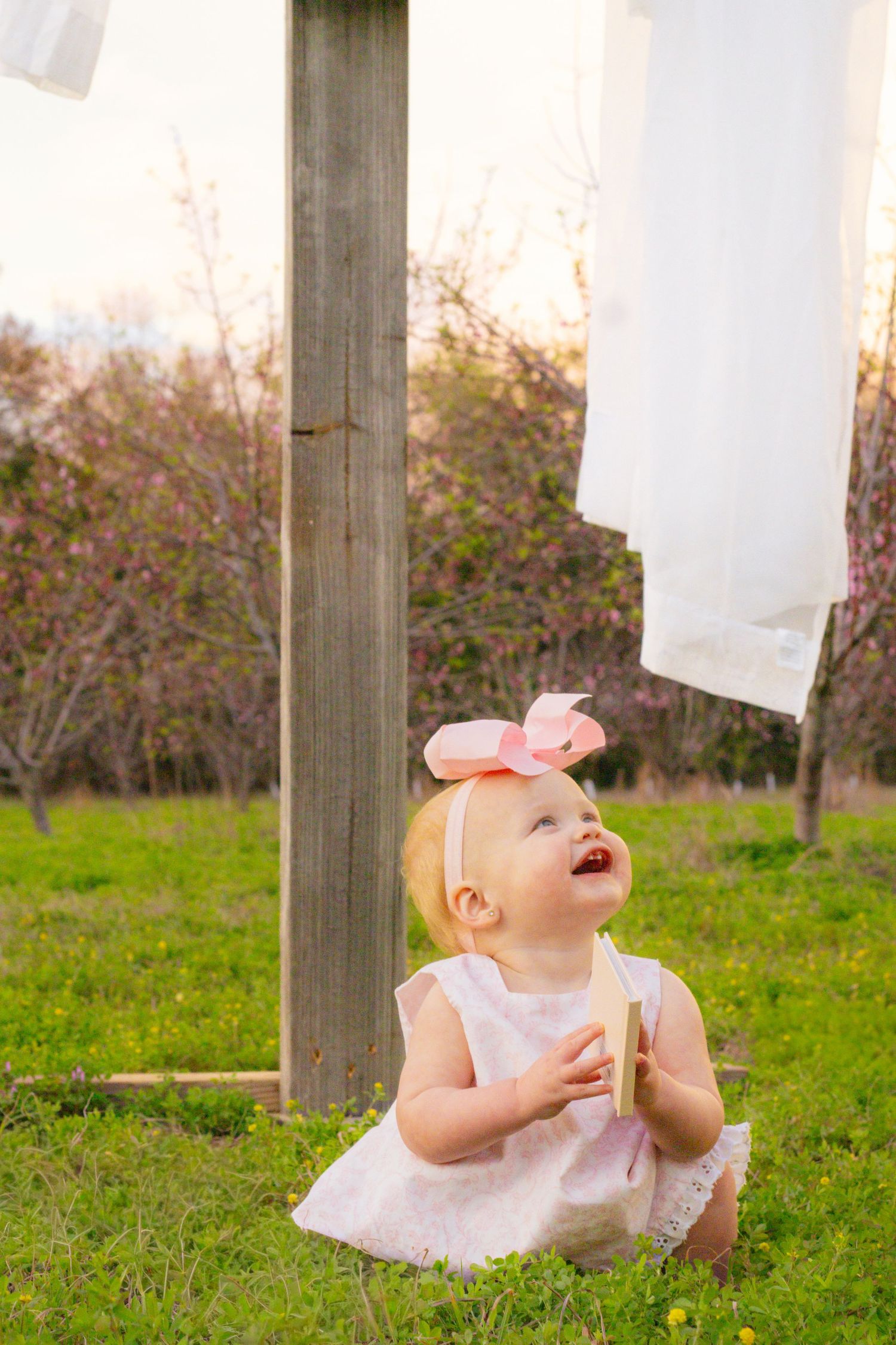 Cute baby girl in pink bow headband sits on grass looking up at white fabric draped on wooden cross.