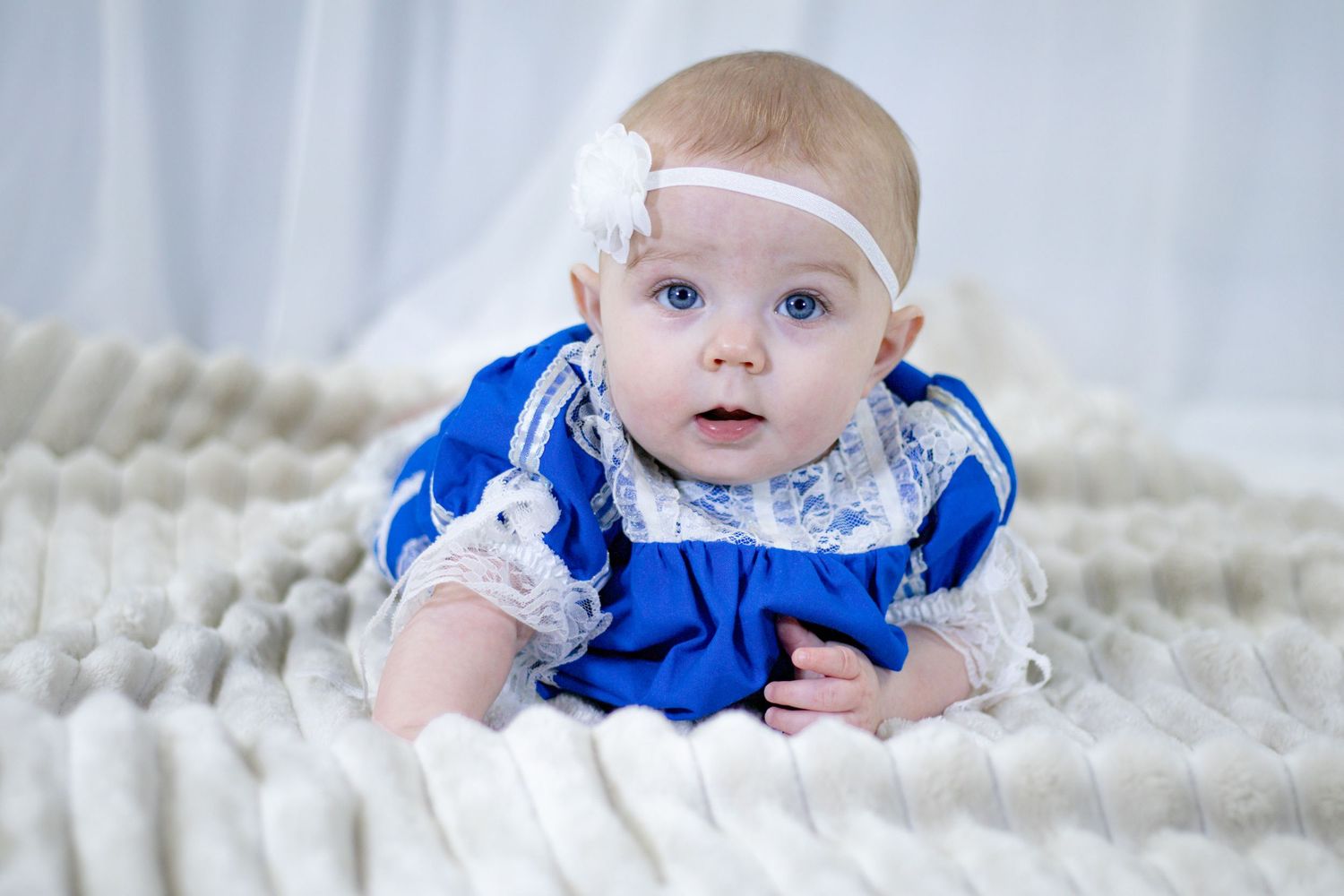Blue-eyed baby girl in blue lace dress and white floral headband doing tummy time on white blanket.