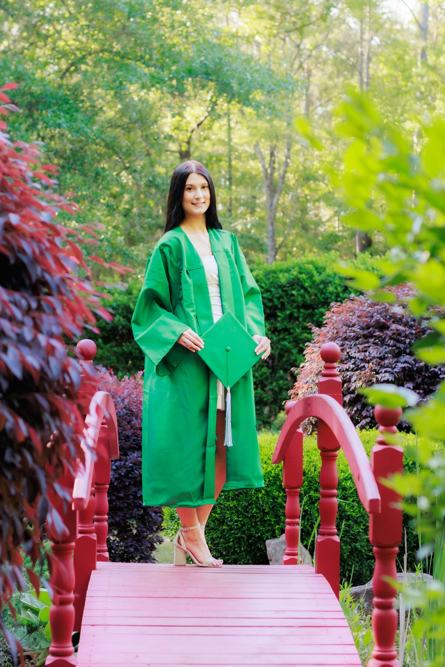 Young woman in green graduation gown and cap standing on a red bridge surrounded by lush garden greenery.