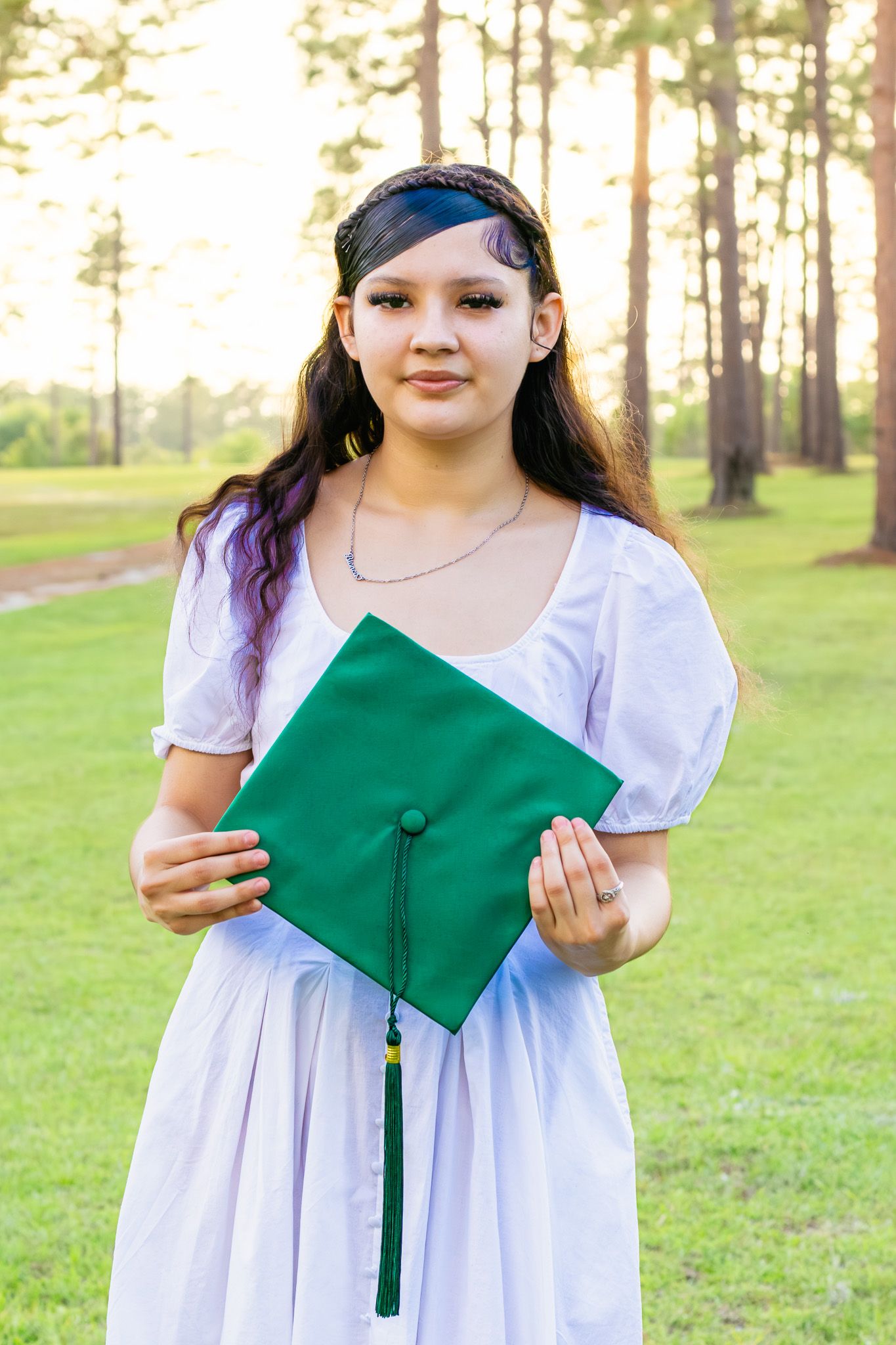 Young woman in white dress holding green graduation cap outdoors among tall pine trees at sunset.