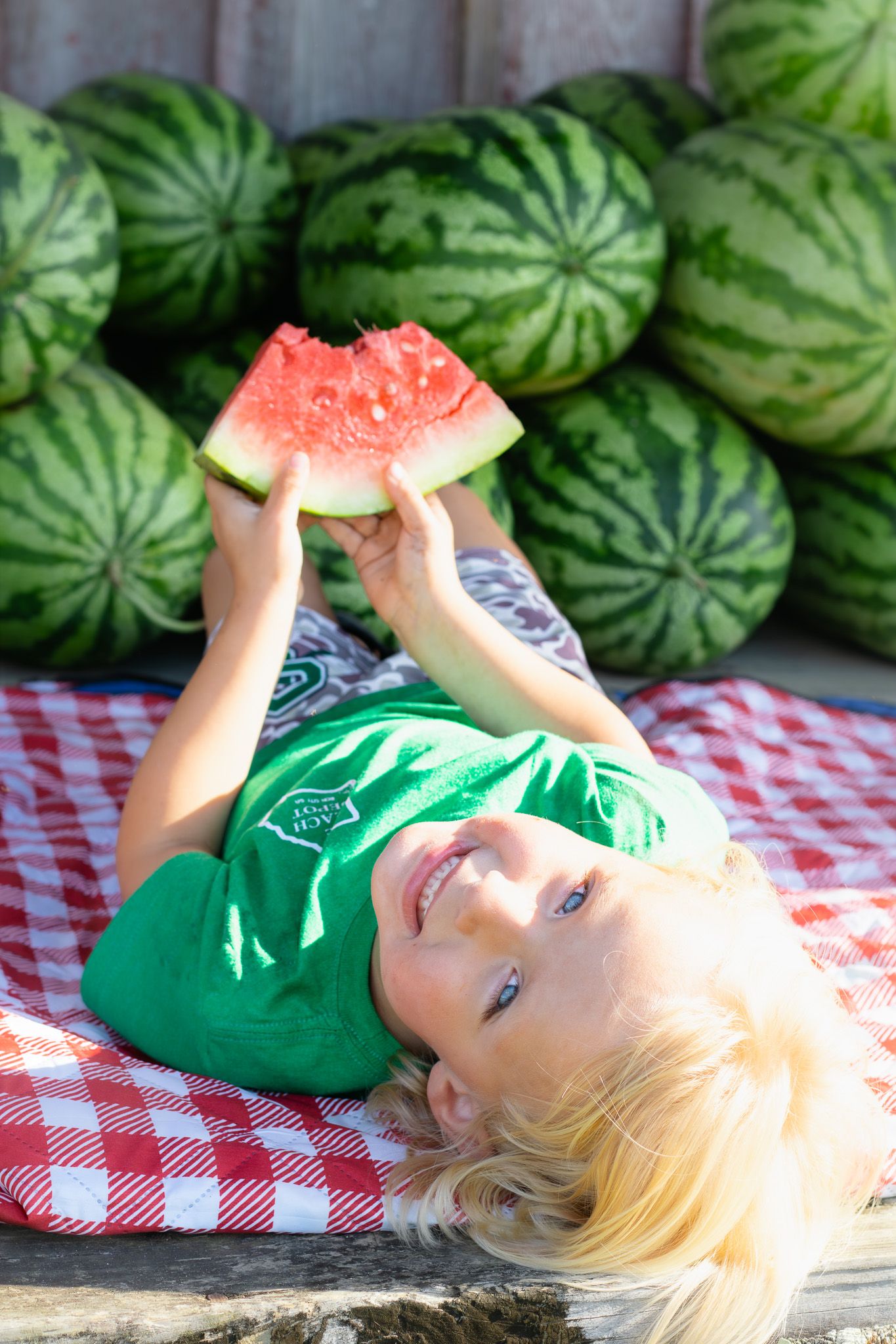 Happy blonde boy lying on red checkered blanket holding up a slice of watermelon with stacked watermelons behind him.