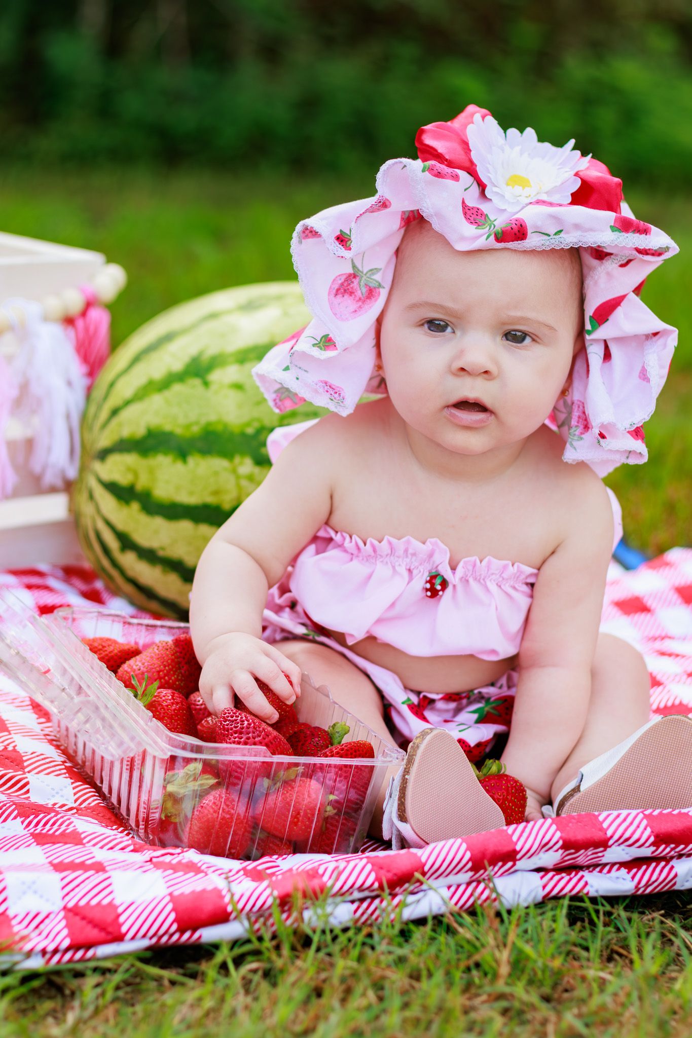 Baby girl in strawberry hat and bikini sits on picnic blanket with fresh strawberries and watermelon outdoors.