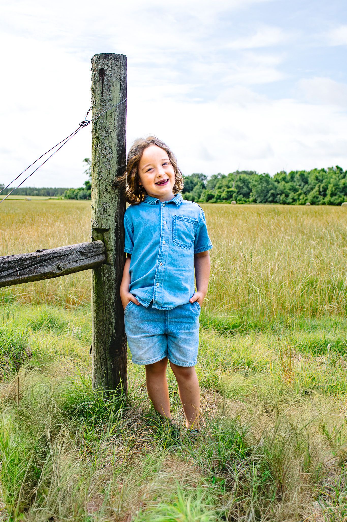 A happy young boy in a blue shirt leans against a wooden fence post in a sunny green field.