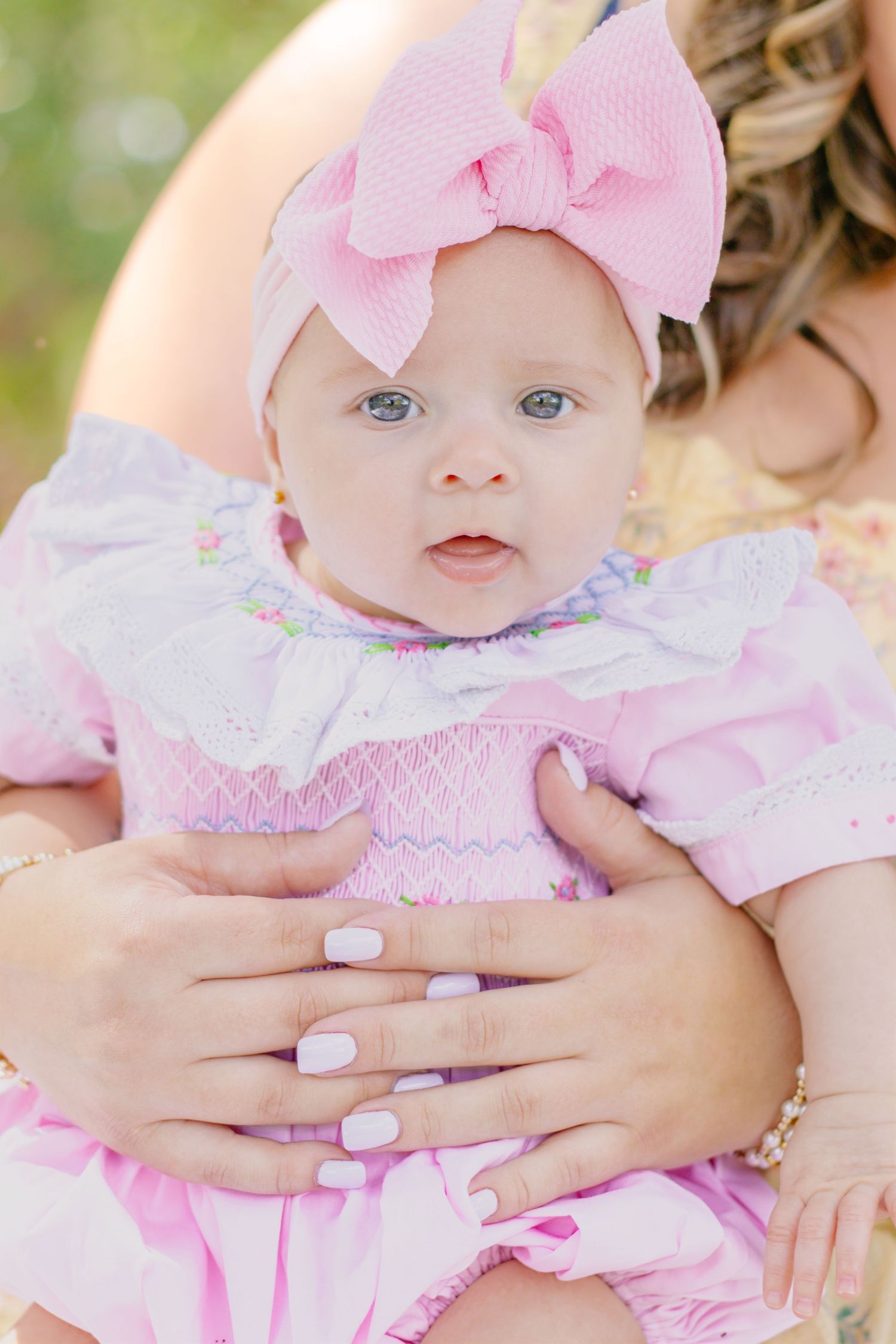 Close-up of blue-eyed baby in pink smocked dress and bow headband cradled in mothers hands.