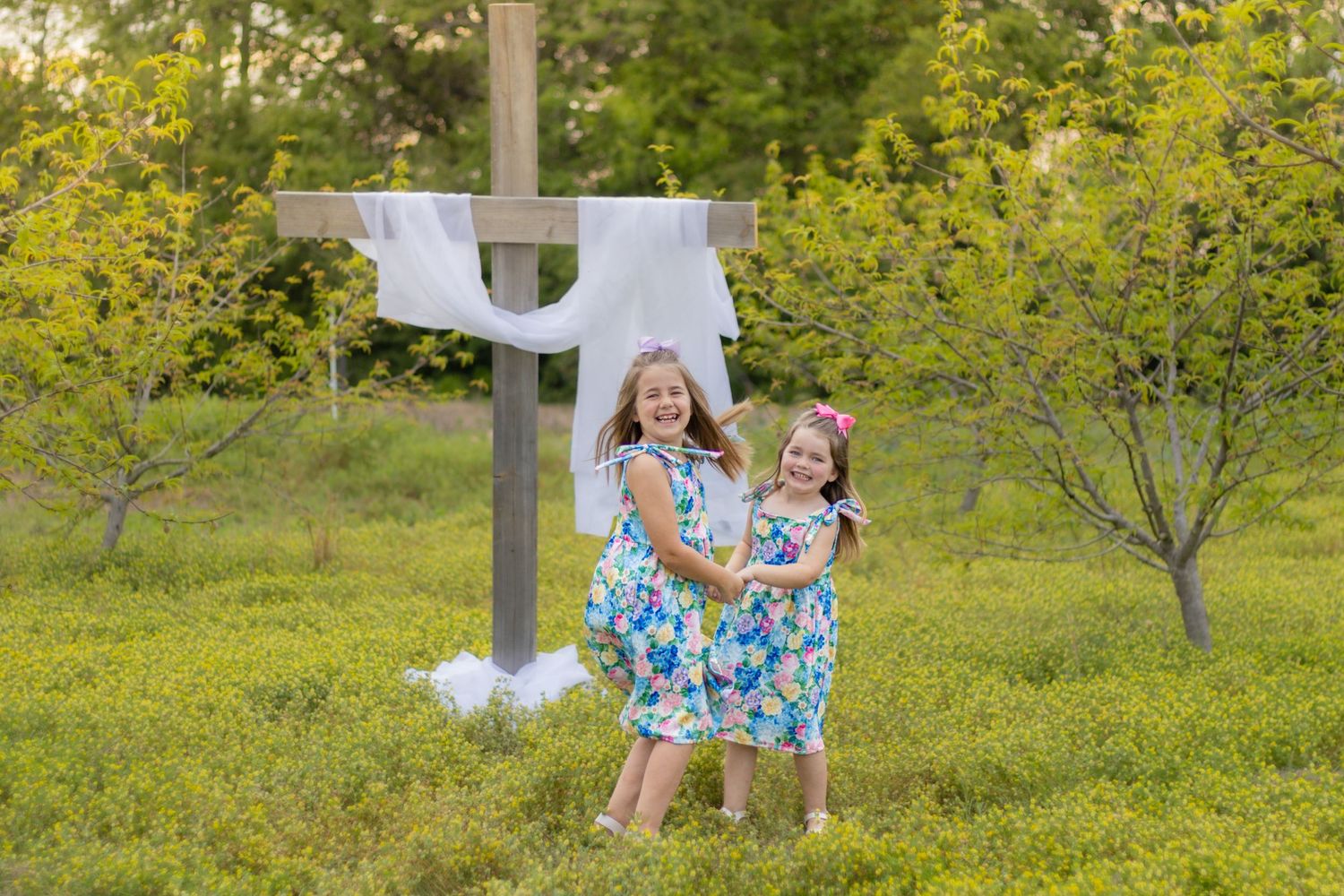 Two young girls in matching floral dresses smiling together in front of a wooden cross draped with white cloth outdoors.