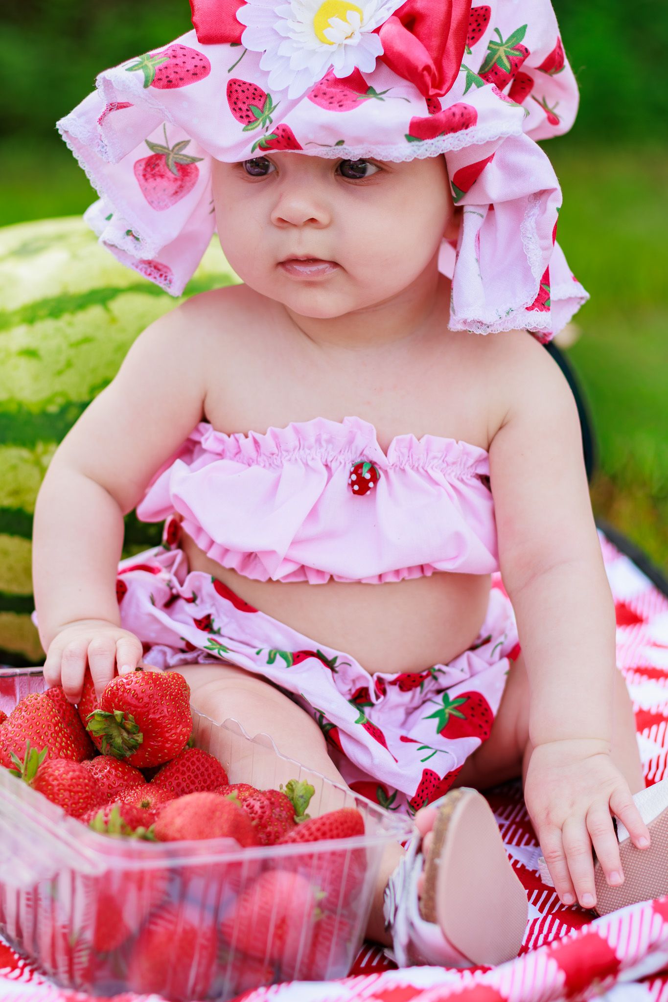 Adorable baby girl in a strawberry-print bikini and sun hat sitting with a container of fresh strawberries outdoors.
