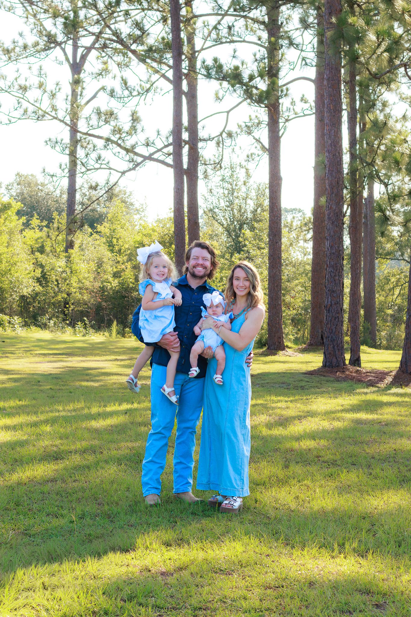 Happy family of four in matching blue outfits posing in a sunlit pine tree meadow.