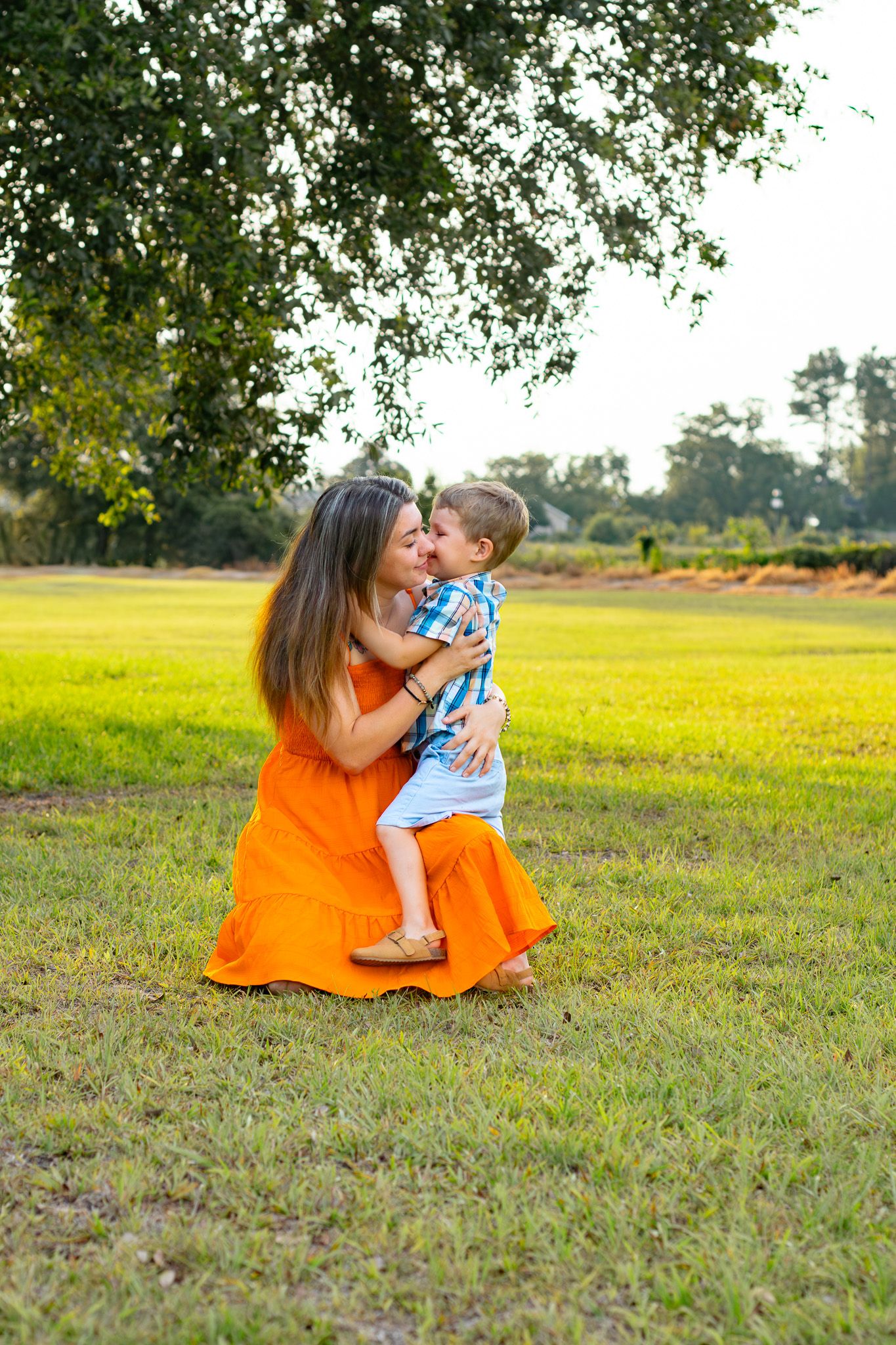 Woman in orange dress kneeling on grass hugging a toddler boy outdoors in a sunny green park.