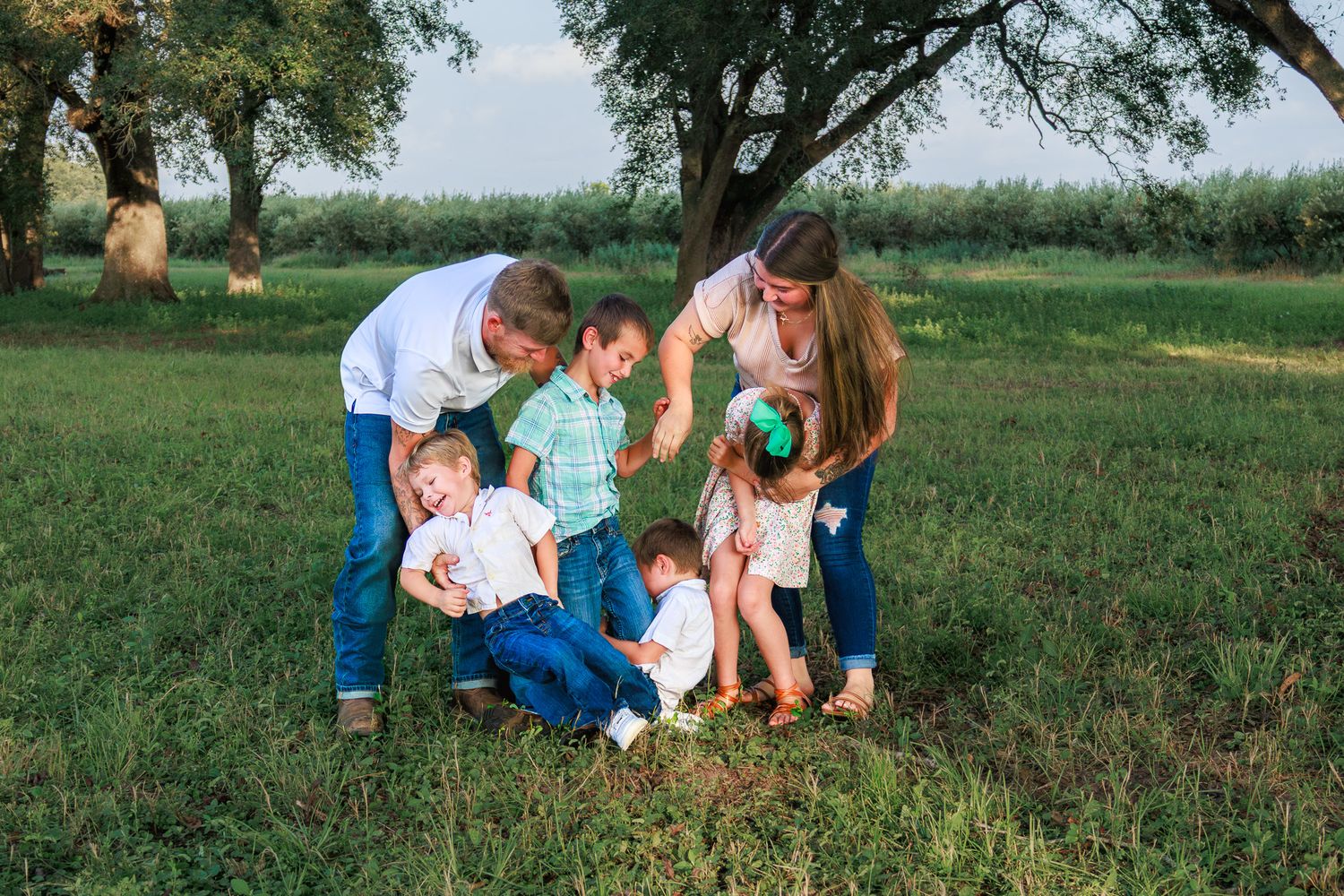 Family of six enjoying outdoor time together on green grass under large trees on a sunny day.