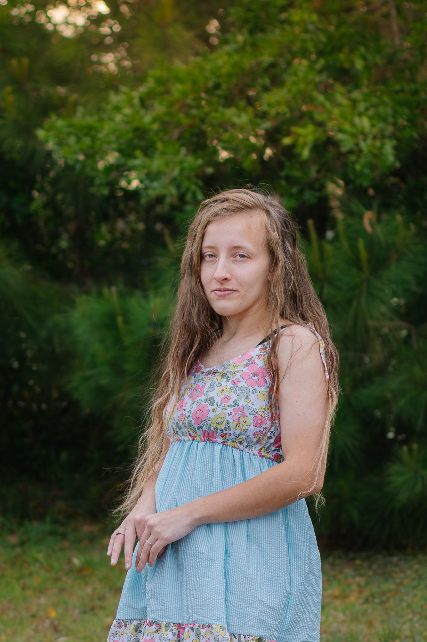 Young girl with long wavy hair poses outdoors in a floral and light blue sundress with green trees behind her.