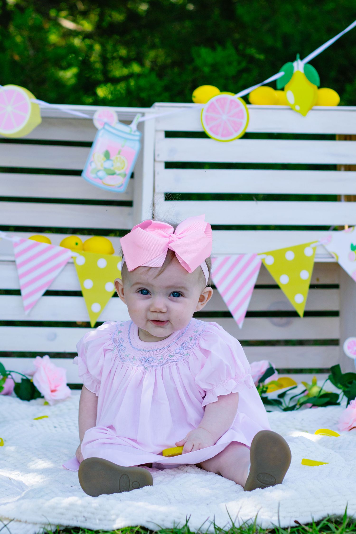 Smiling baby girl in pink smocked dress with bow headband at lemon-themed outdoor birthday setup.