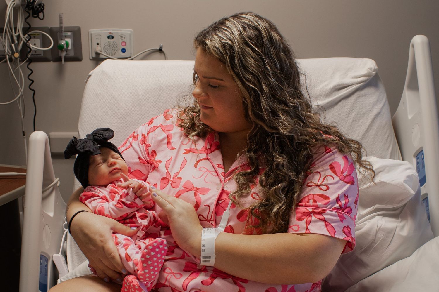 New mother in hospital bed lovingly holding her newborn baby girl in pink outfit.