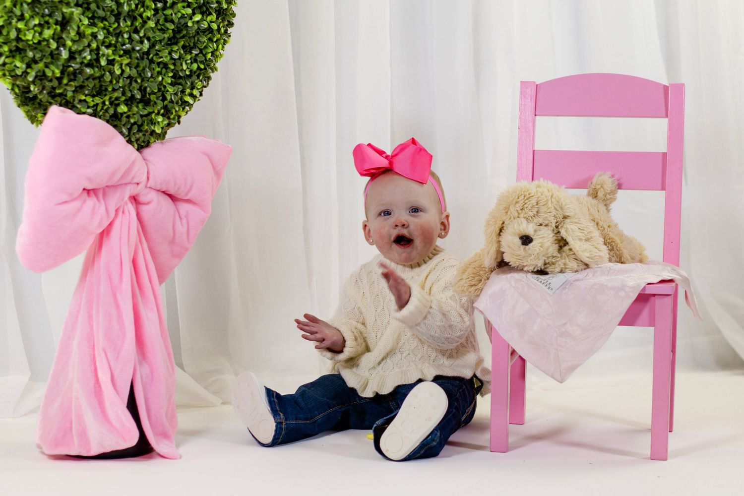 Adorable baby girl in cream sweater holds a stuffed animal beside a pink chair and large decorative bow.