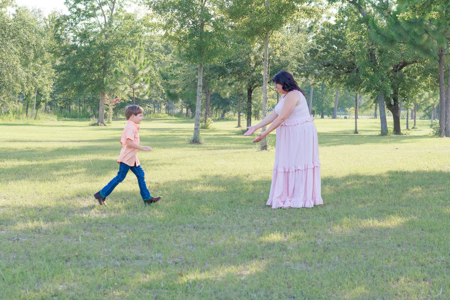 A woman in a pink dress reaches out to a young boy running toward her in a lush green park.