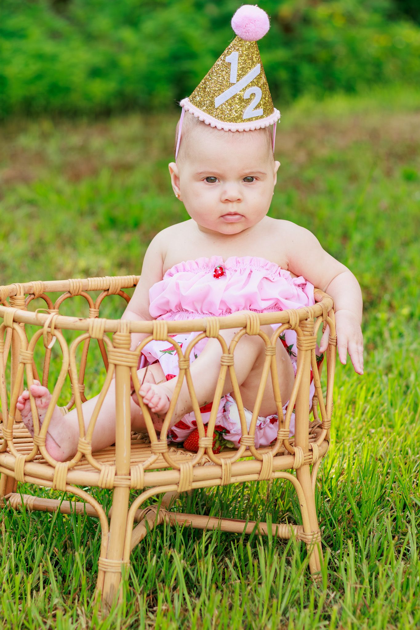 Baby girl in pink outfit and gold half birthday hat sits in wicker chair on green grass outdoors.