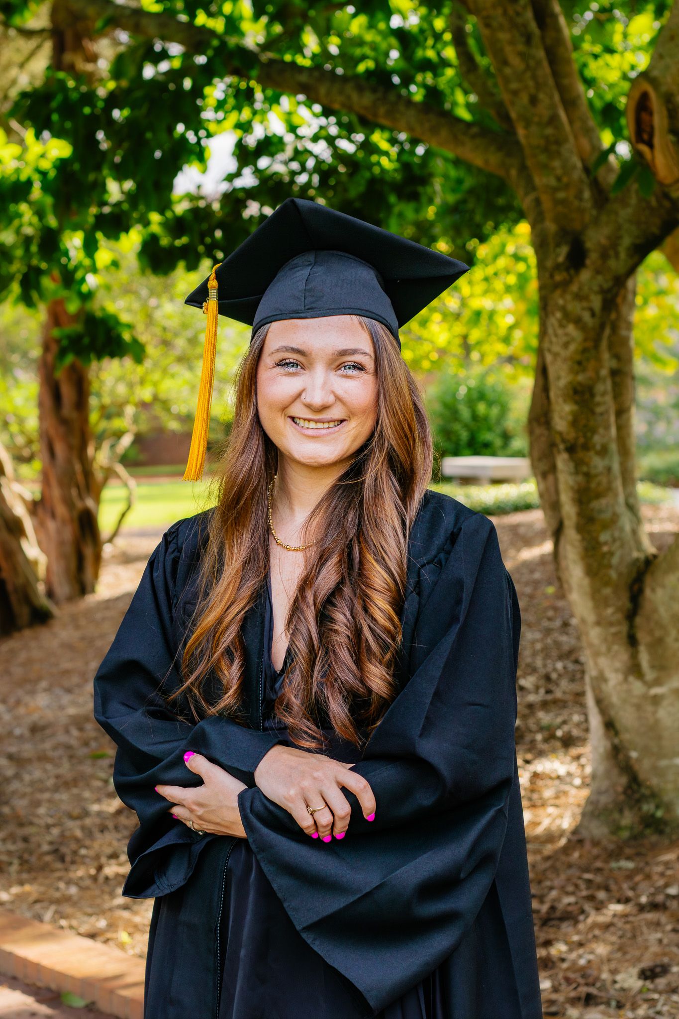 Smiling graduate in black cap and gown poses outdoors among green trees.