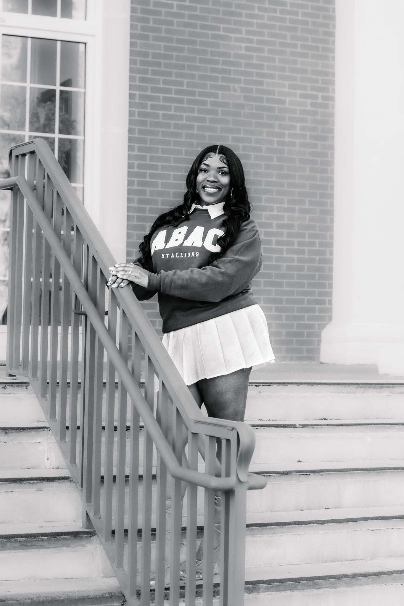 Young woman in ABAC Stallions sweatshirt and pleated skirt posing on brick building staircase in black and white photo.