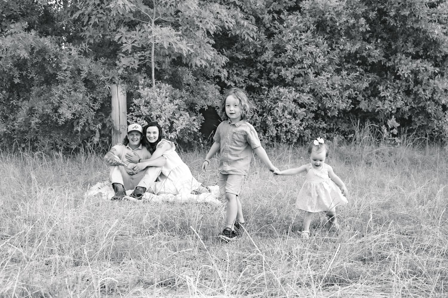 Black and white photo of a family relaxing and playing in a meadow with tall grass and trees behind them.