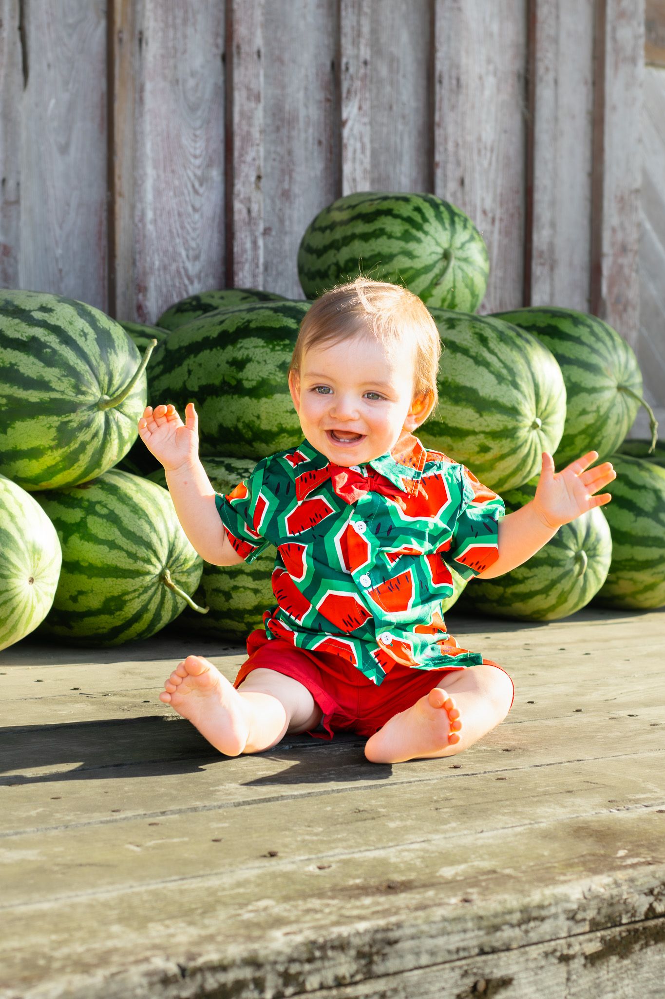 Happy baby in watermelon print shirt sits barefoot on wooden deck surrounded by large green watermelons.