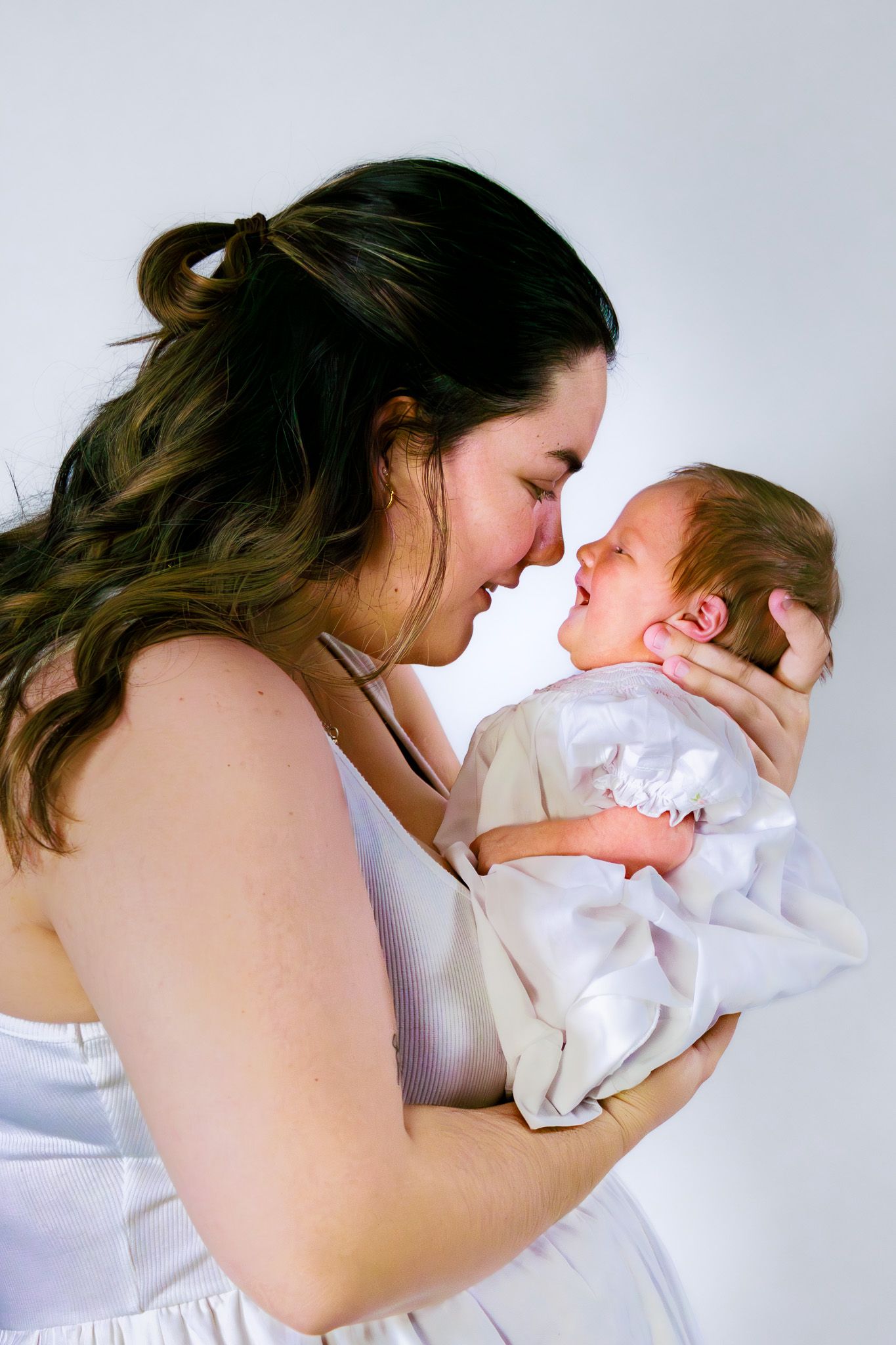 Mother smiling and touching noses with her newborn baby wrapped in white blanket.