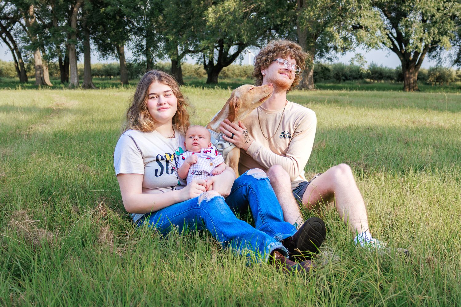Young couple sitting in green grass field holding a newborn baby, smiling outdoors with trees in background.