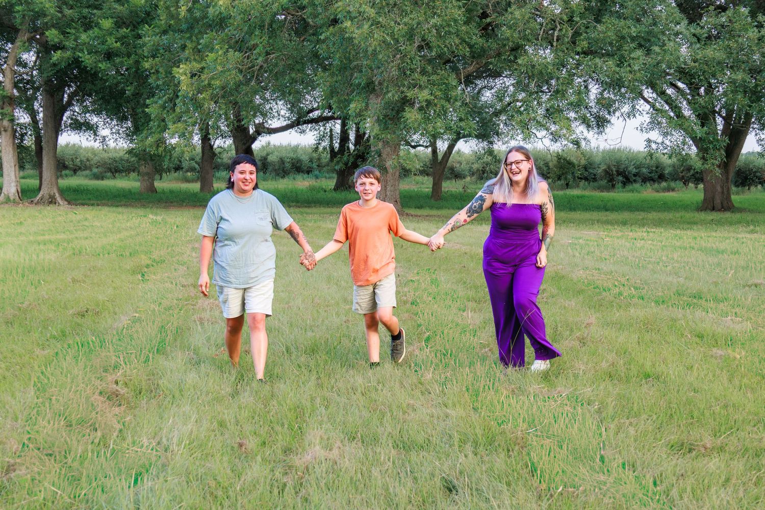 Family of three holding hands walking through a lush green field surrounded by trees on a summer day.