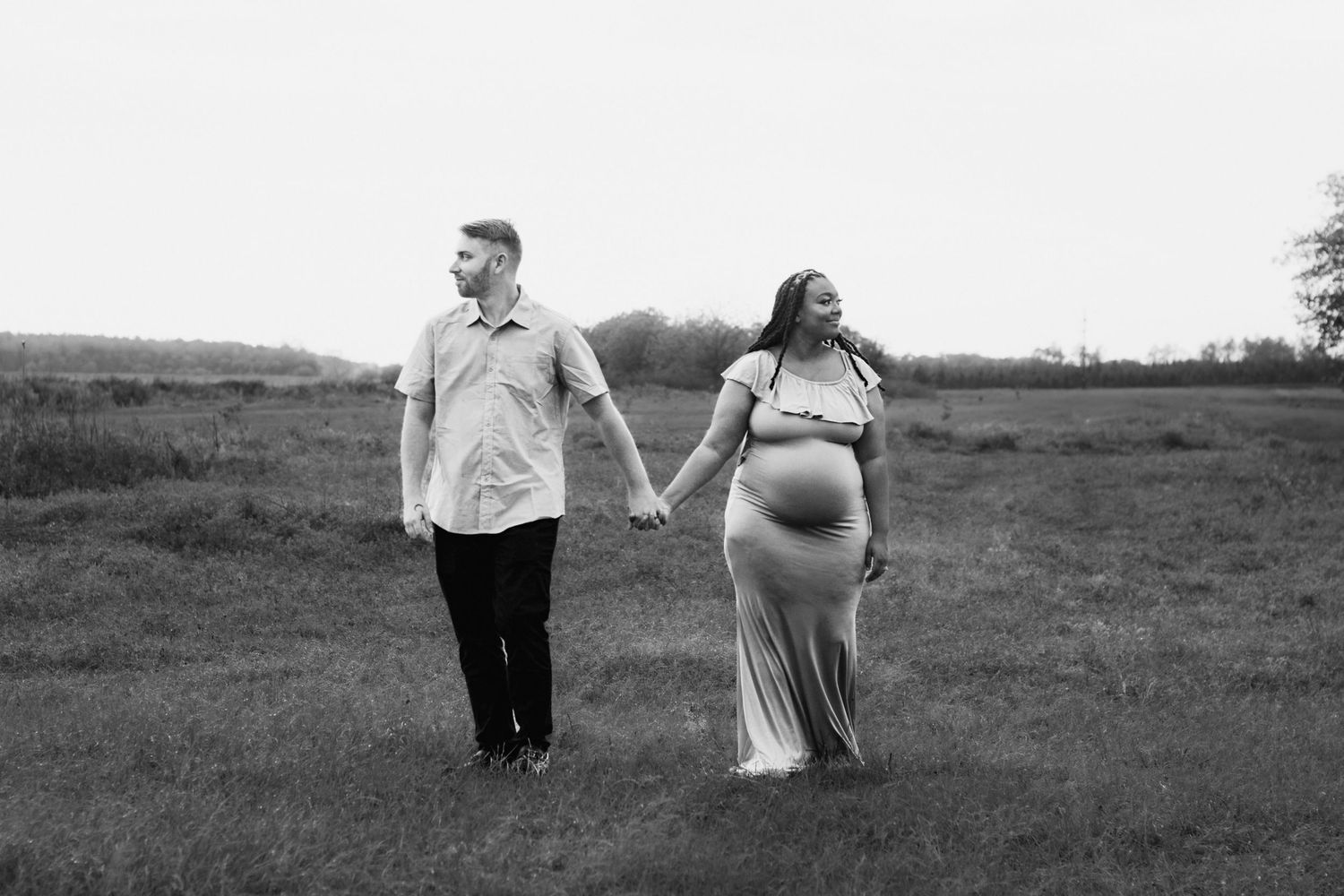 Black and white photo of a pregnant couple holding hands walking through a green field during maternity photoshoot.
