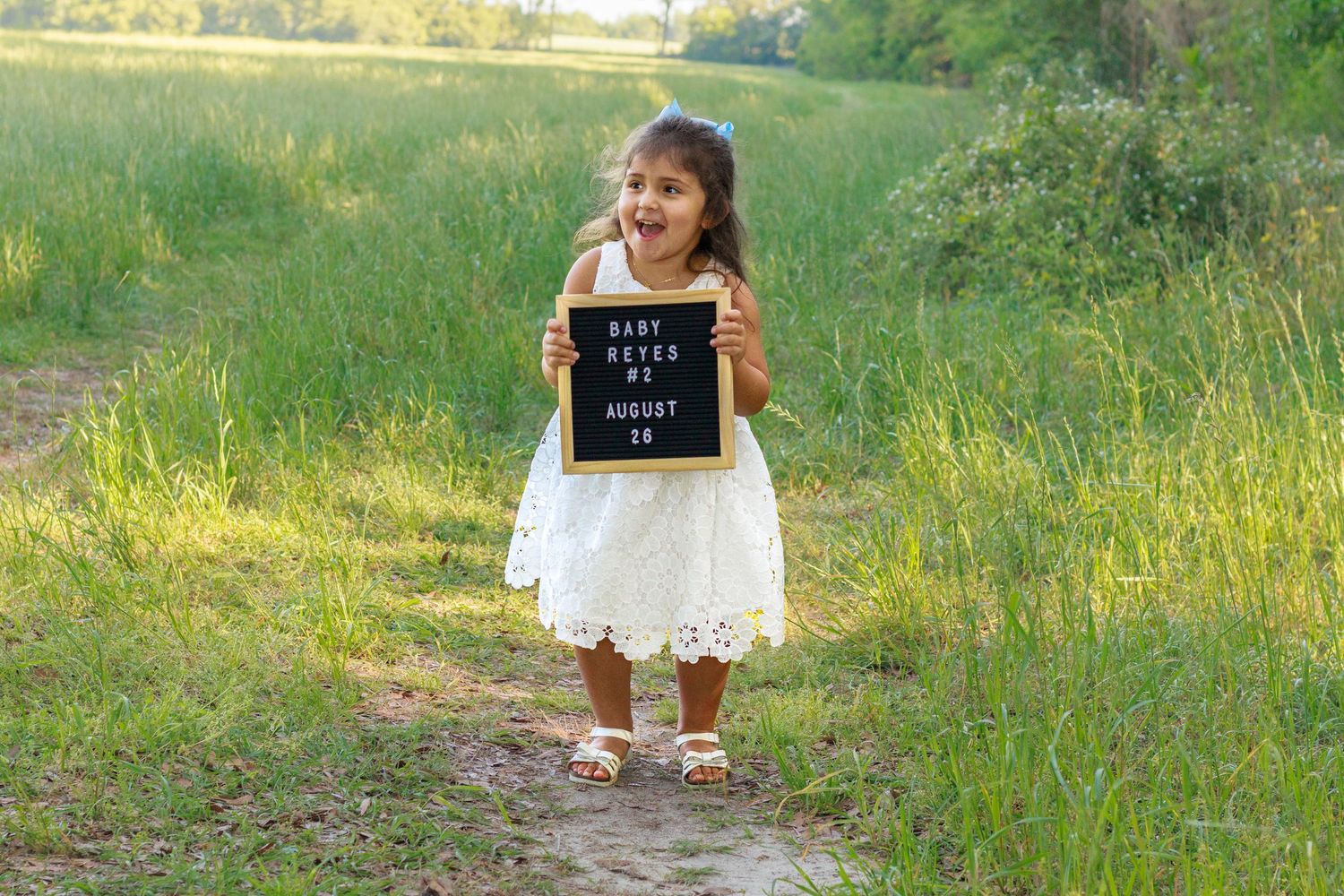 Young girl in white dress holding a letter board announcing Baby Reyes #2 due August 26.