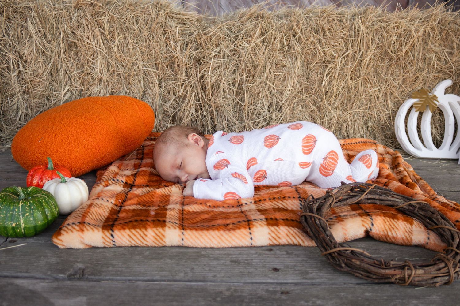 Swaddled newborn sleeping peacefully on plaid blanket surrounded by pumpkins and hay bales.