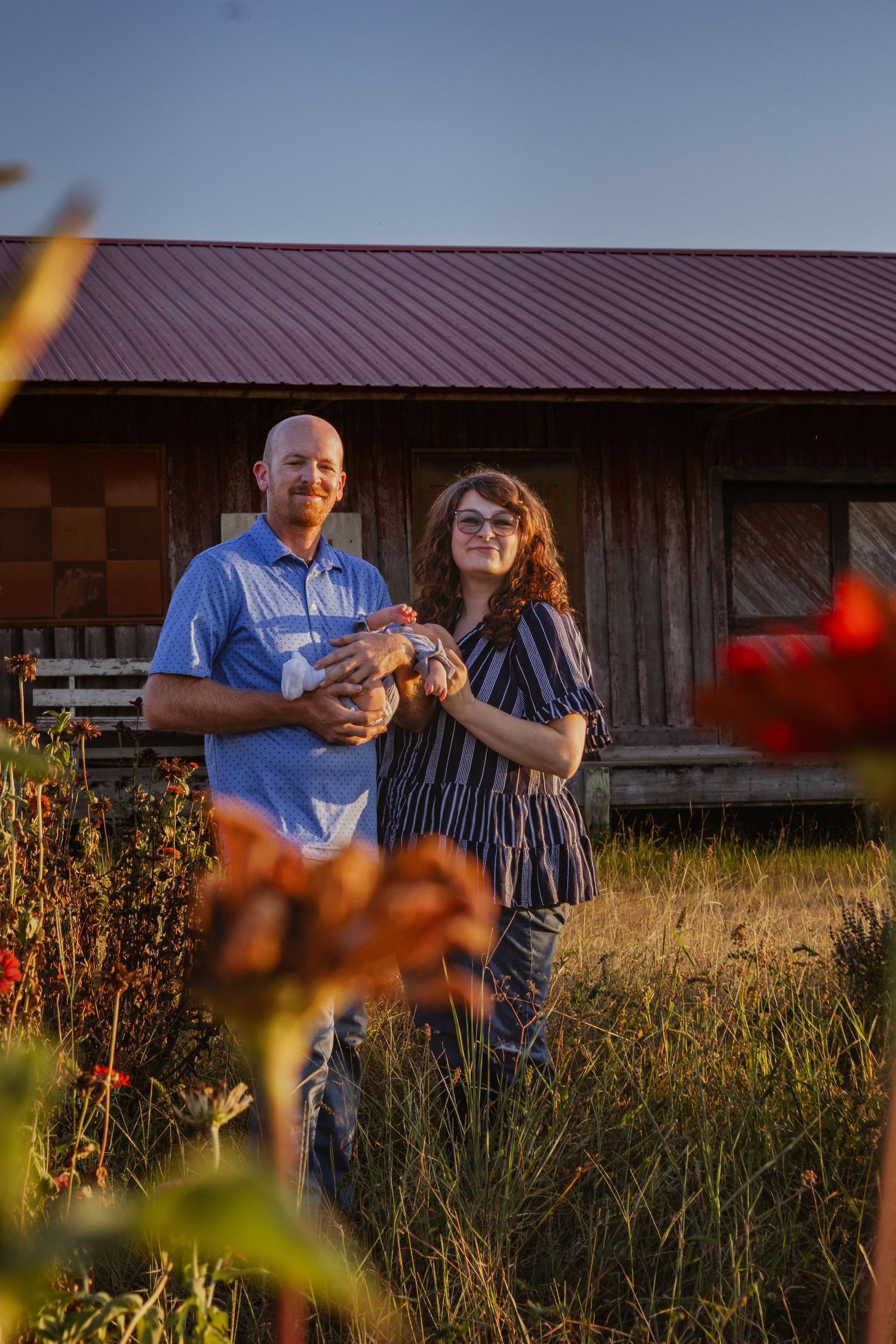 Couple smiling and holding a chicken in front of a rustic barn surrounded by wildflowers at golden hour.