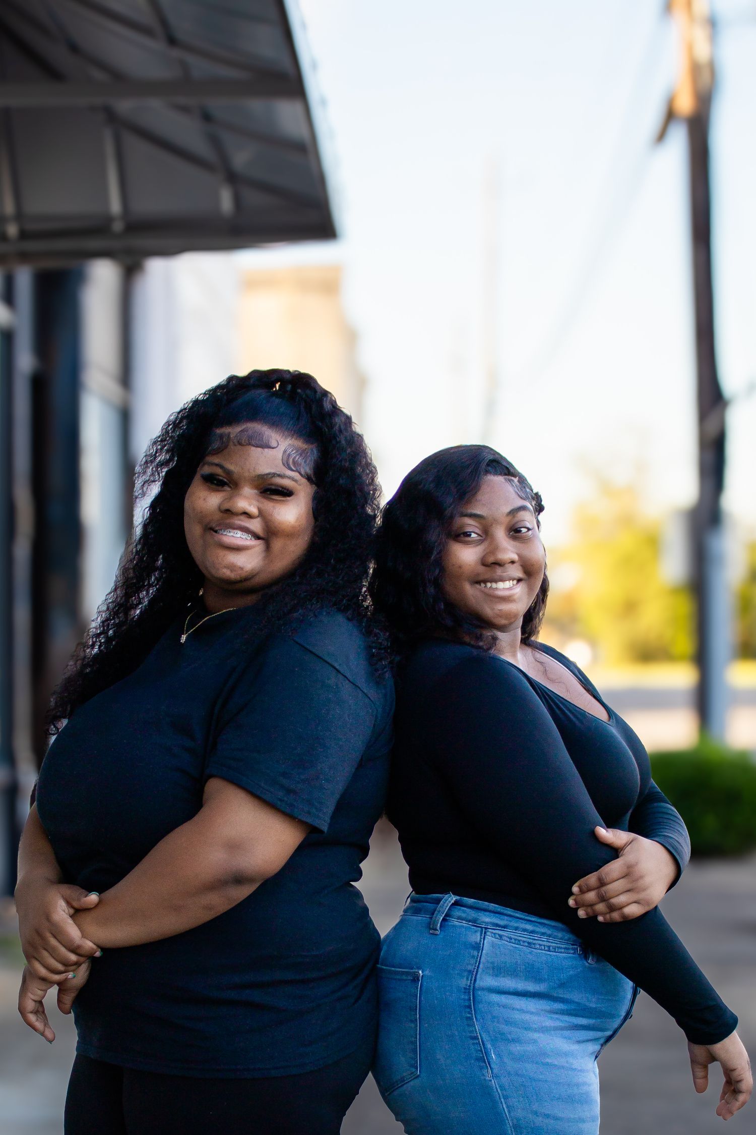 Two smiling young Black women in black tops posing back-to-back on a sunny urban sidewalk.