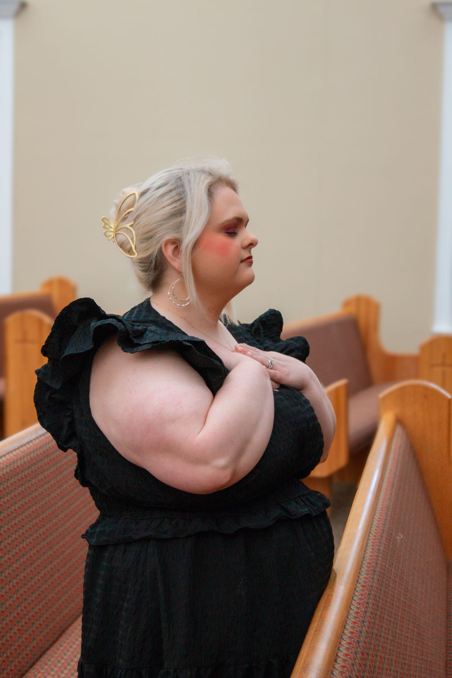 A plus-size woman in a black ruffled dress prays reverently in church pews with gold hair accessories.